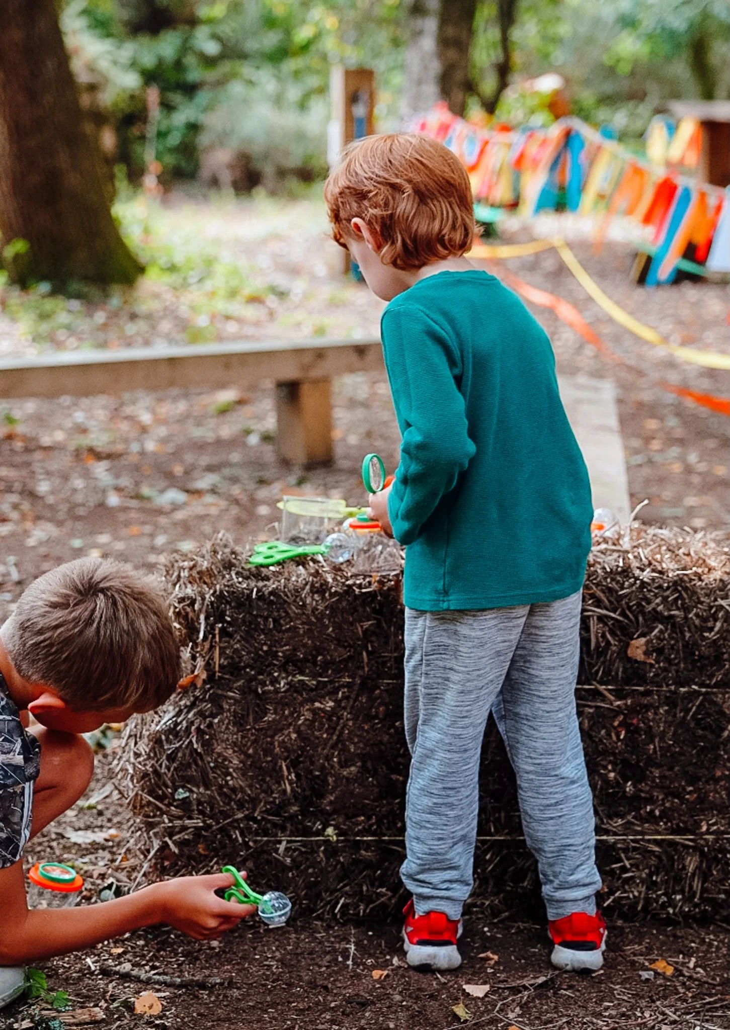 Two young boys playing outdoors, digging and exploring in a wooded area. One boy is standing with gray sweatpants and red sneakers, while the other boy is crouching with a toy in hand. The background features trees and a colorful fence.