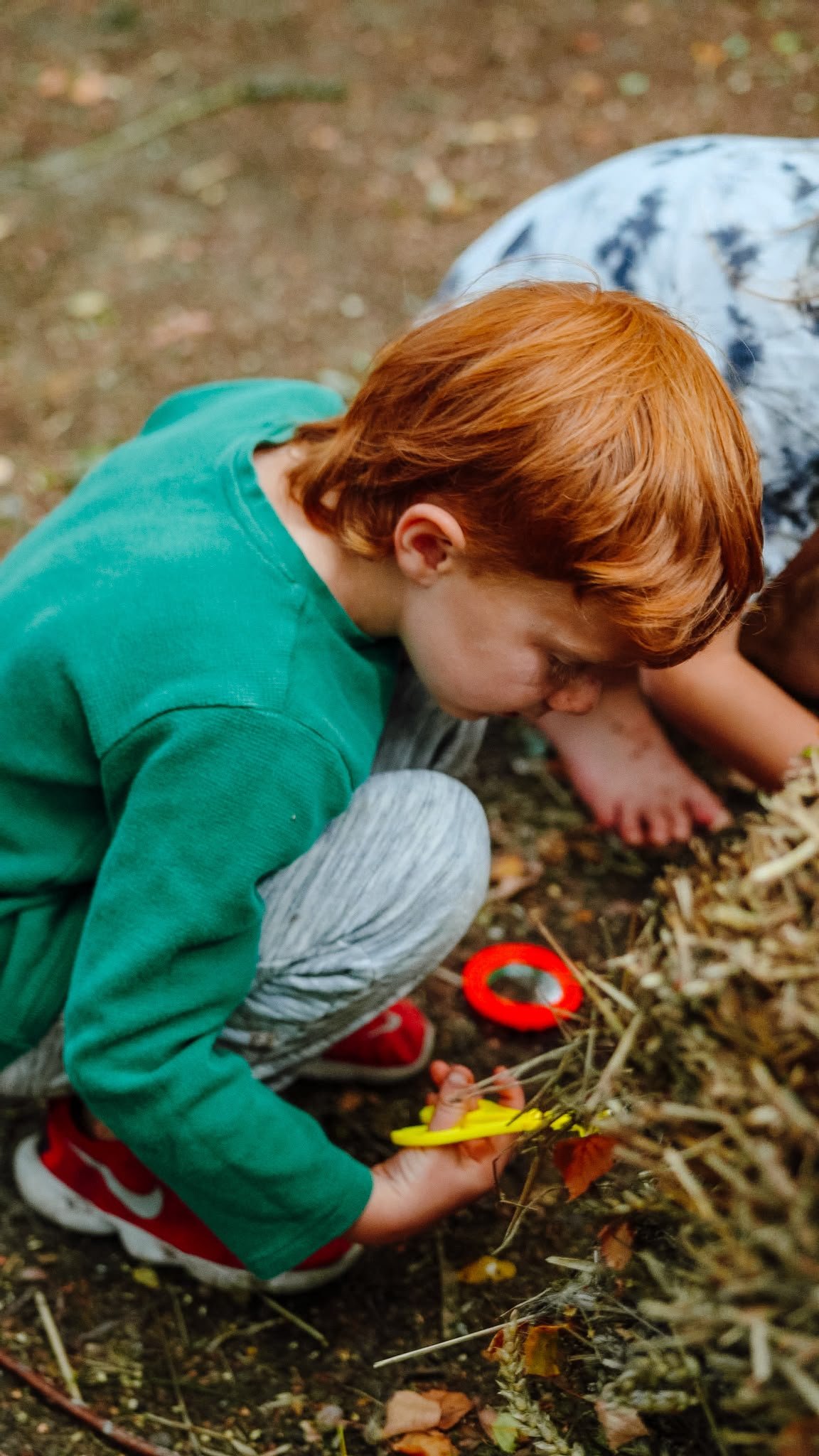A woman and a young girl smiling and looking at a smartphone outdoors during a camping trip, with trees and other people in the background.