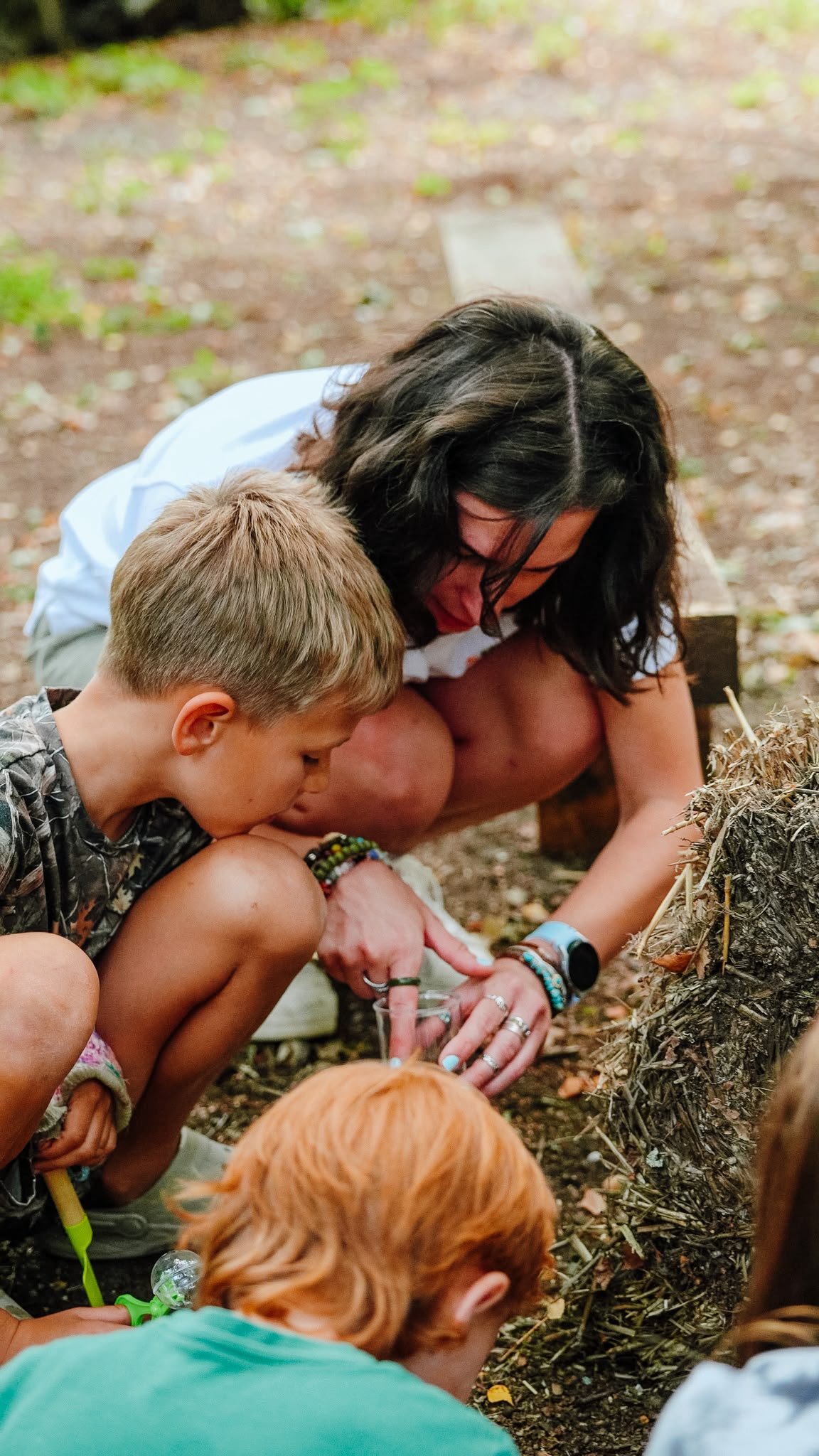 A woman and children crouch down outdoors, engaged in an activity involving soil or plants, with trees and leaves in the background.