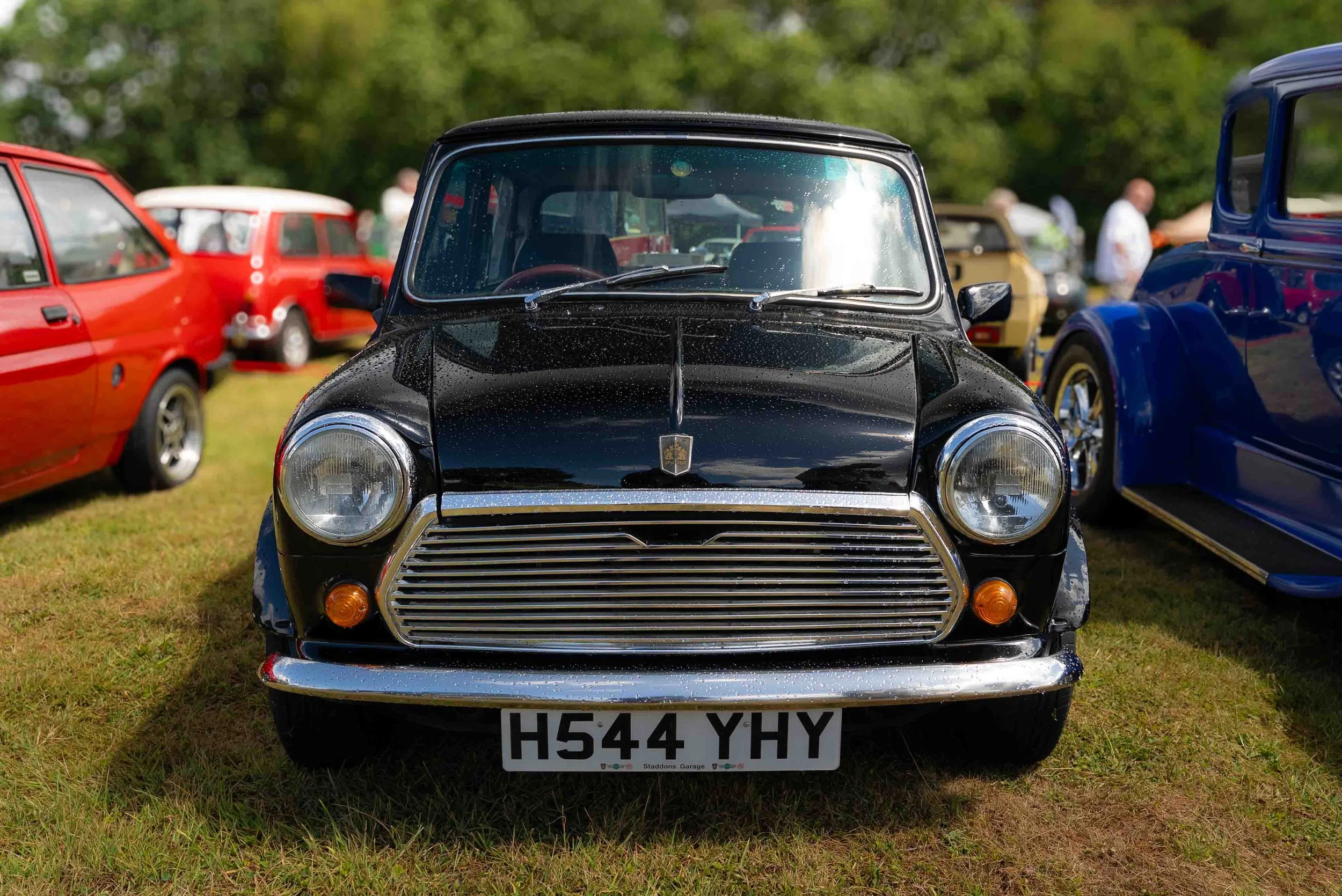 Front view of a black vintage Mini Cooper car with water droplets, parked on grass at a car show, surrounded by other classic cars and people.