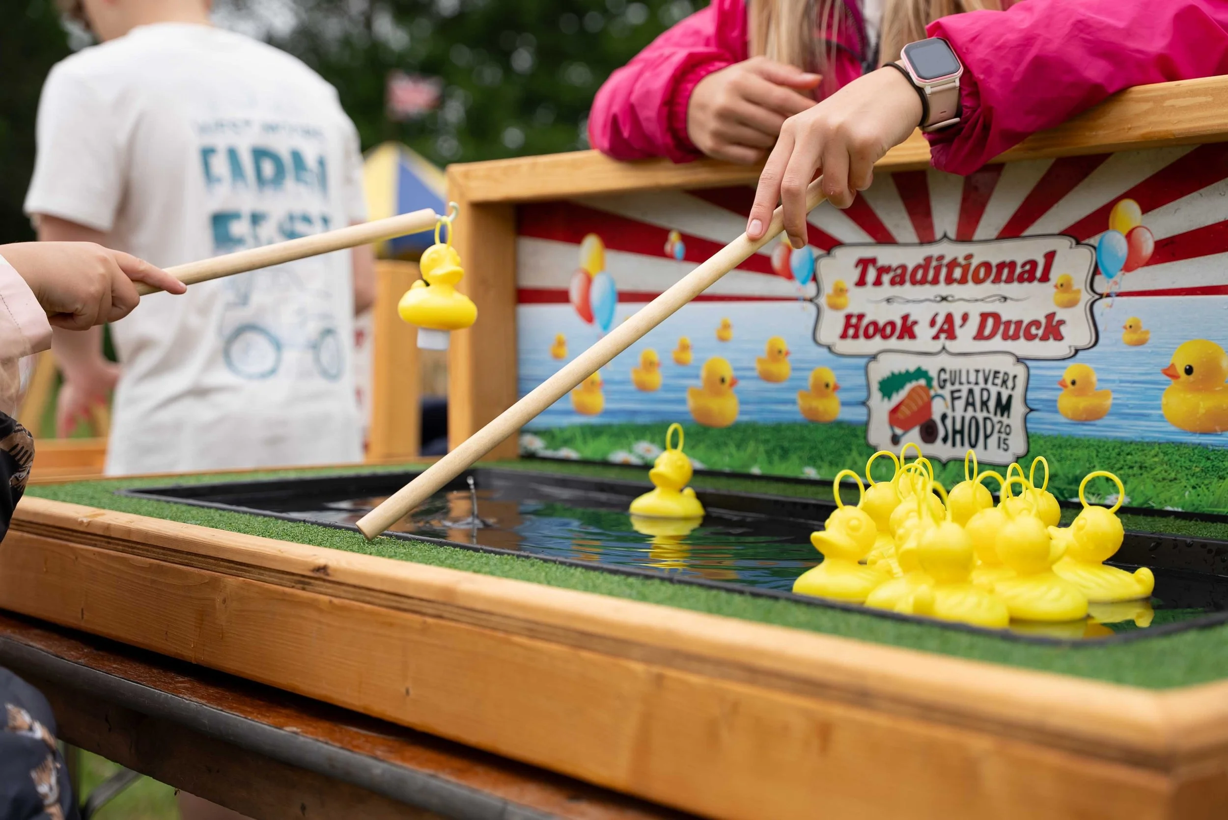 People playing a duck pond fishing game at an outdoor fair, with a sign reading 'Traditional Hook A' Duck' and multiple yellow rubber ducks floating in the water.