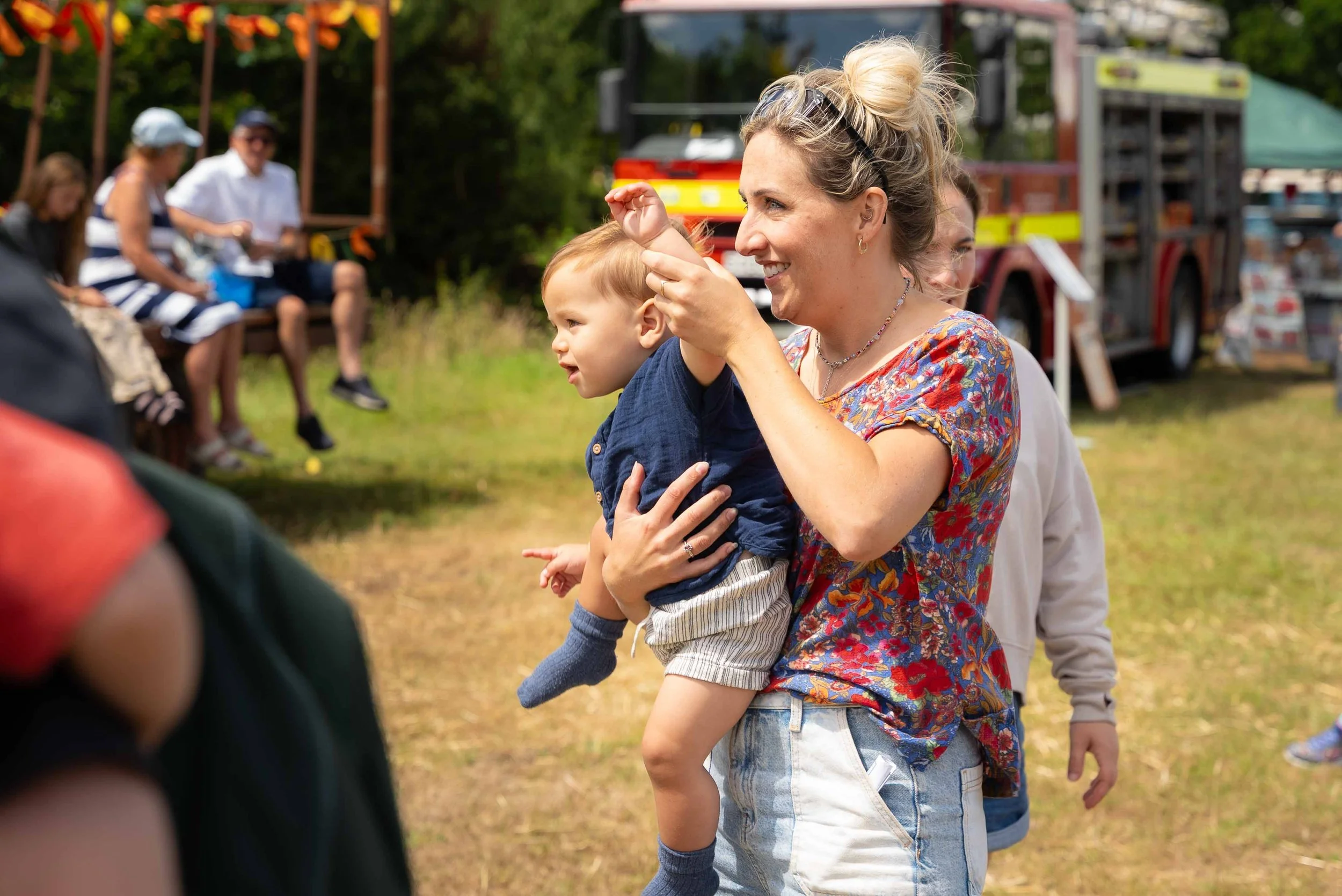 A woman holding a young boy at an outdoor event, with people sitting on benches and a firetruck in the background.