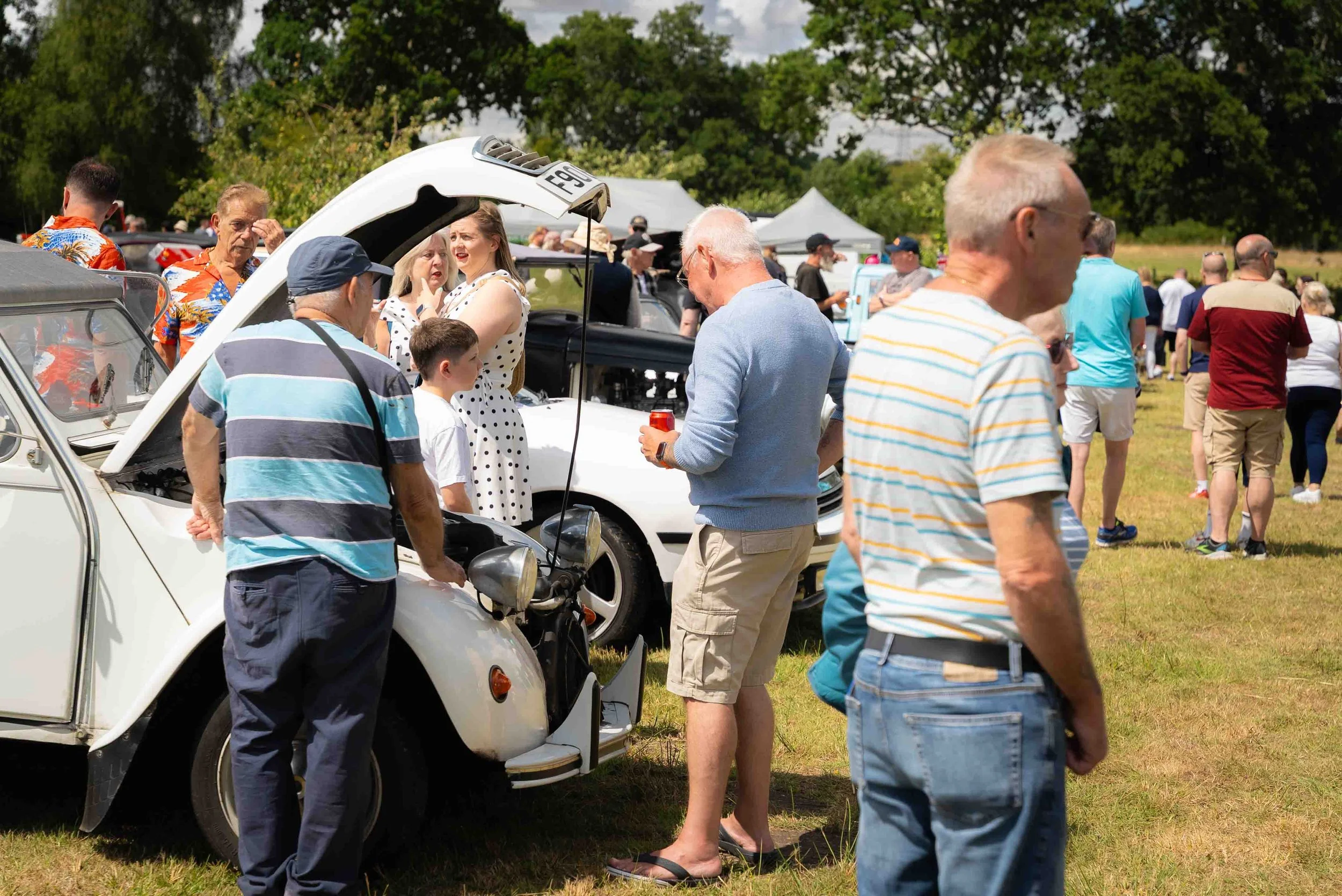People gather at an outdoor car show on a grassy field, looking at classic and vintage cars, with trees and tents in the background.