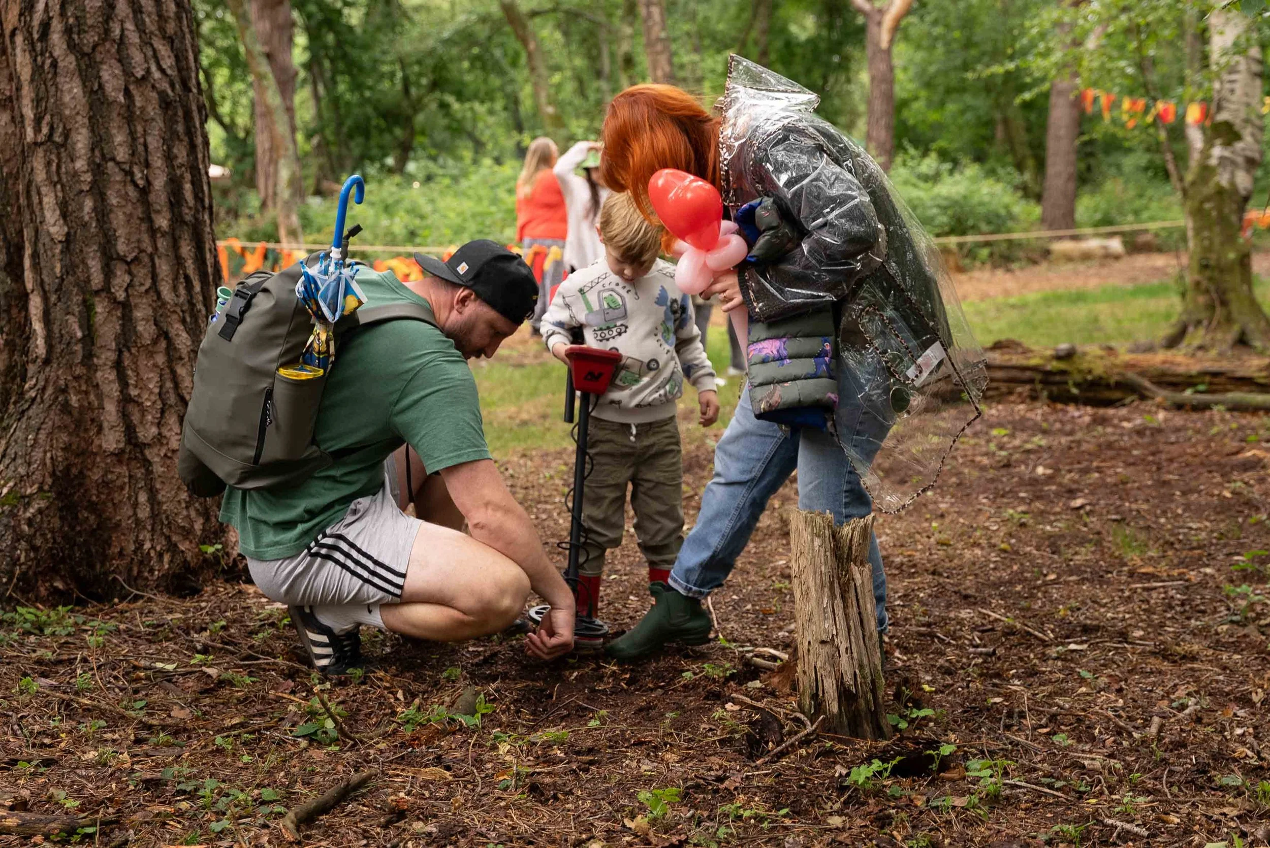 A man, woman, and child in a forest, with the man crouched down looking at the ground, the woman holding balloons and pointing to something, and the child holding a metal detector.