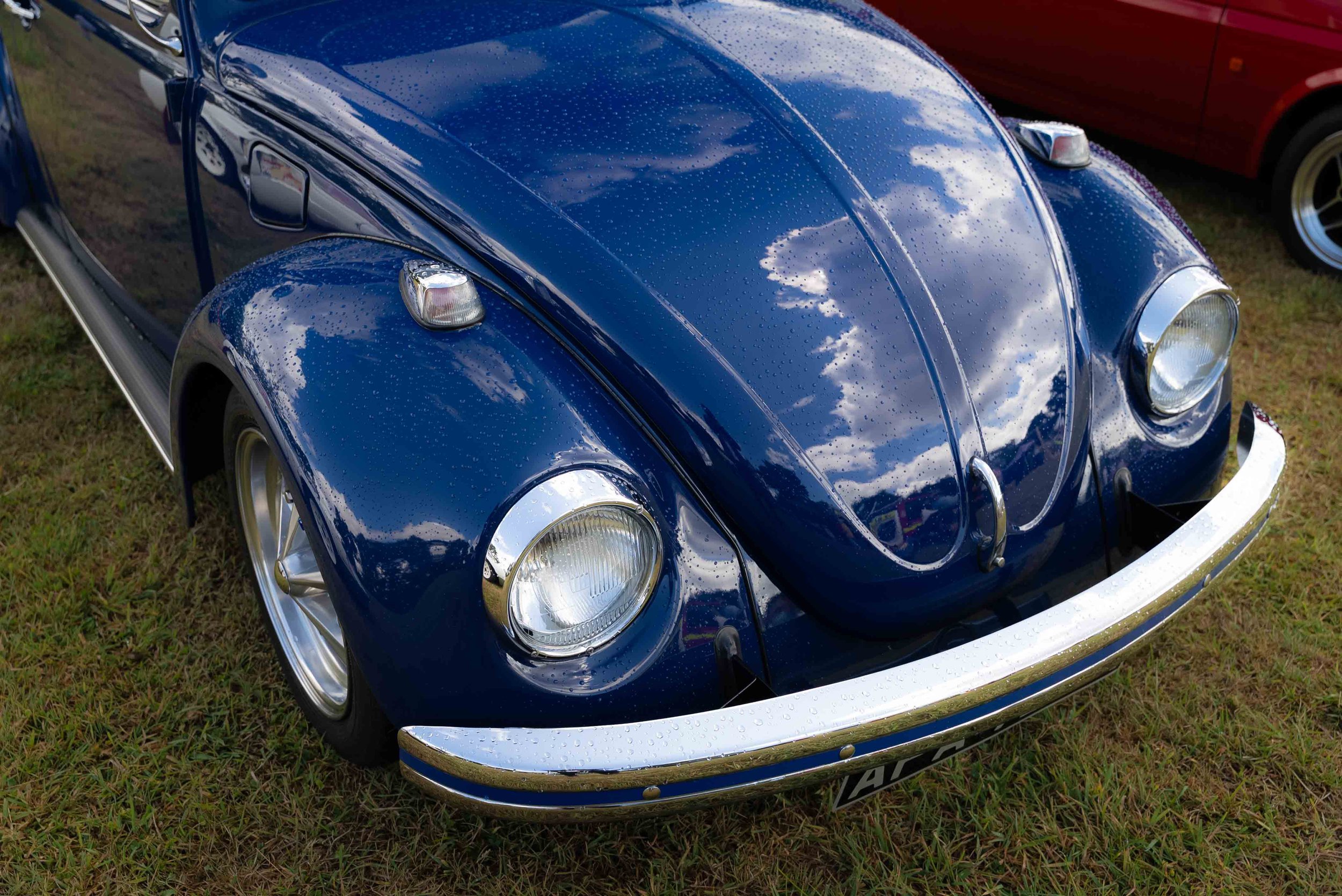 A vintage blue Volkswagen Beetle car with raindrops on its surface, parked on grass.