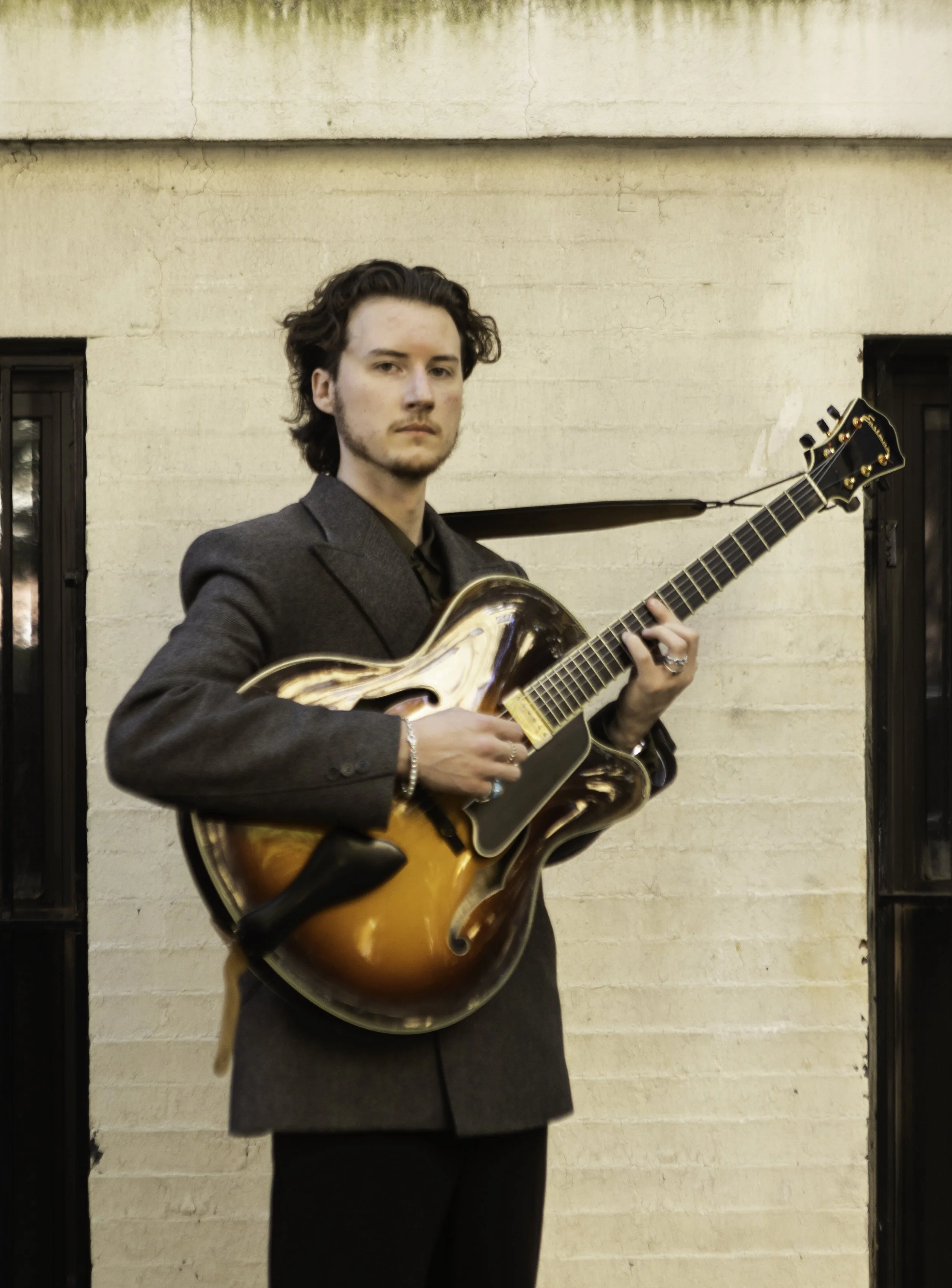A young man with wavy dark hair wearing a gray blazer and black pants plays an orange and brown hollow-body electric guitar outdoors against a beige brick wall.