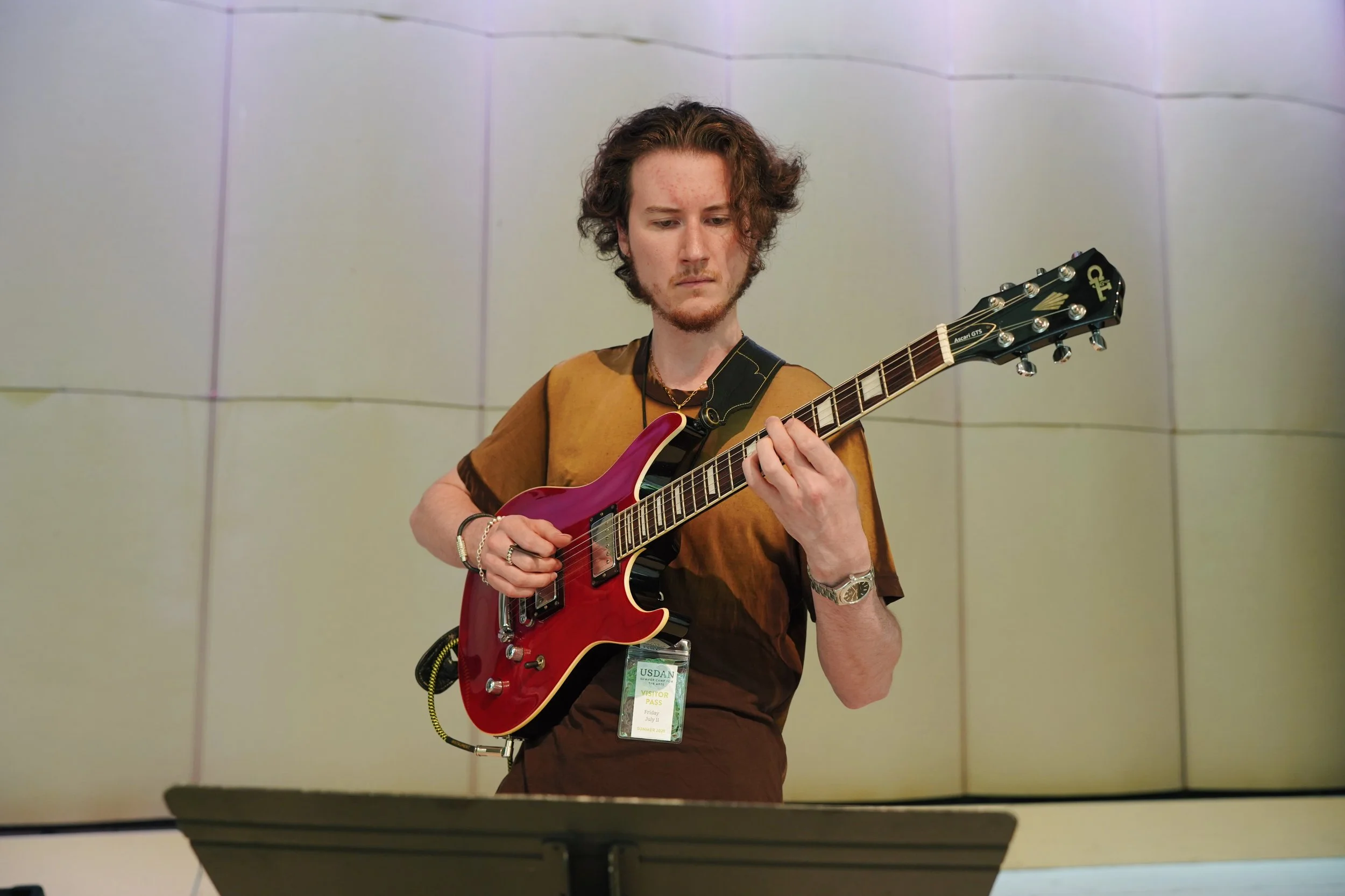 A young man with curly brown hair and freckles playing an electric guitar in a room with beige walls.