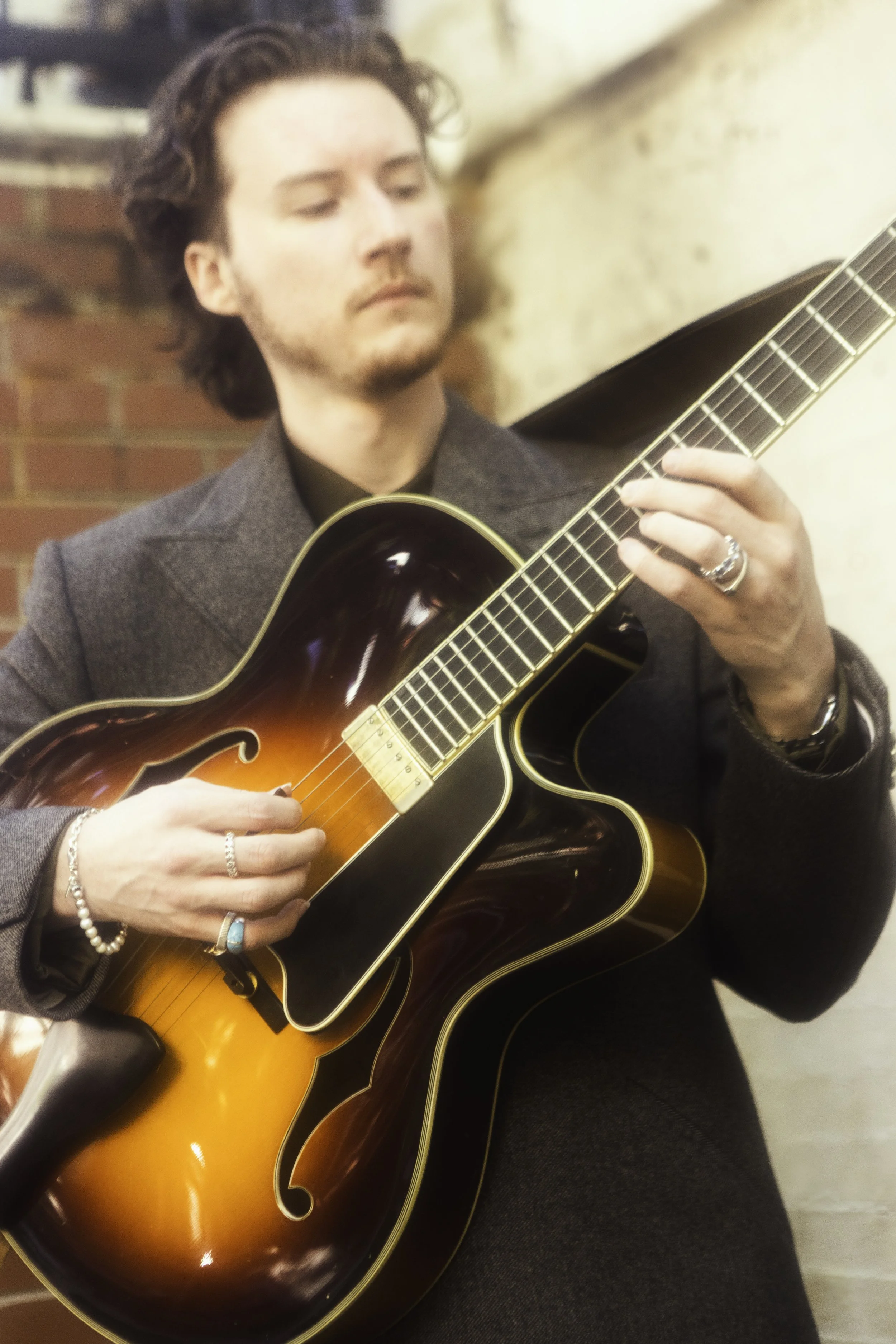 A man with dark wavy hair playing an acoustic guitar with a sunburst finish, wearing a dark blazer and multiple rings.