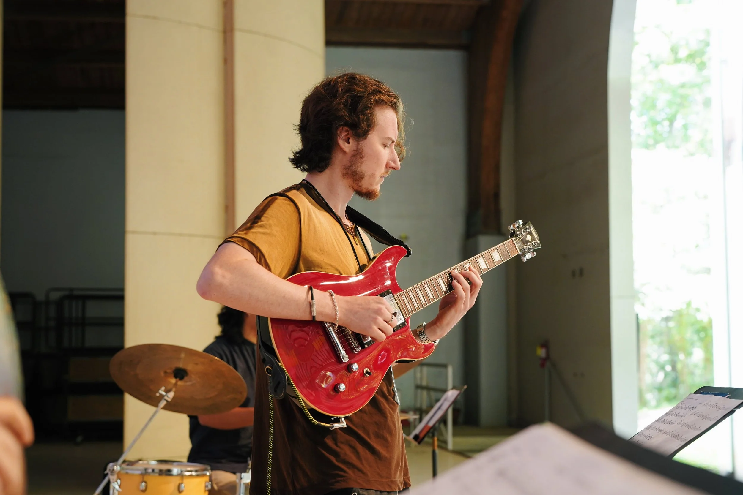 Person playing a red electric guitar in a studio with a drummer in the background and music stand with sheet music nearby.