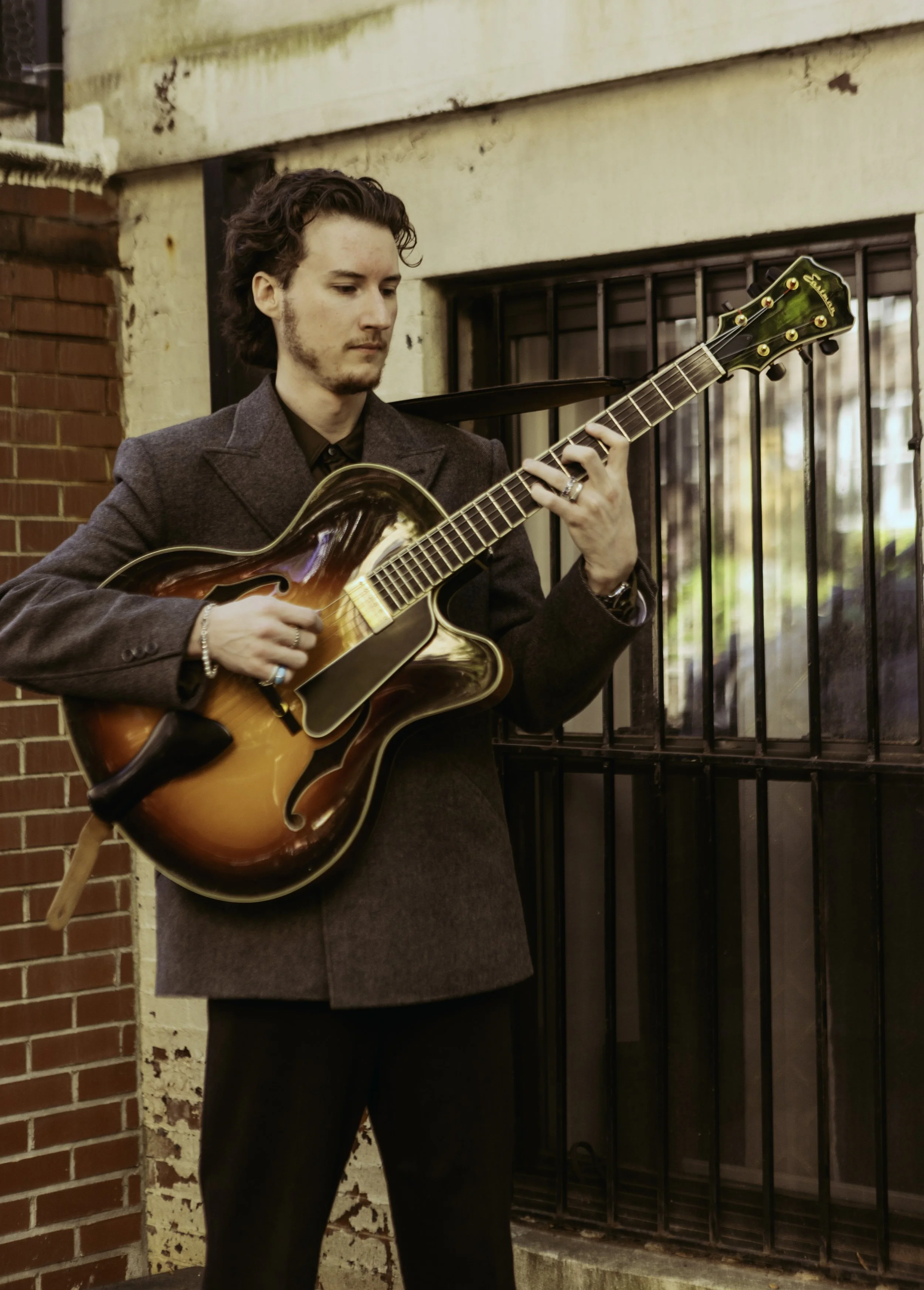 A young man with dark curly hair and light skin playing an electric guitar outdoors near a brick wall and metal gate.