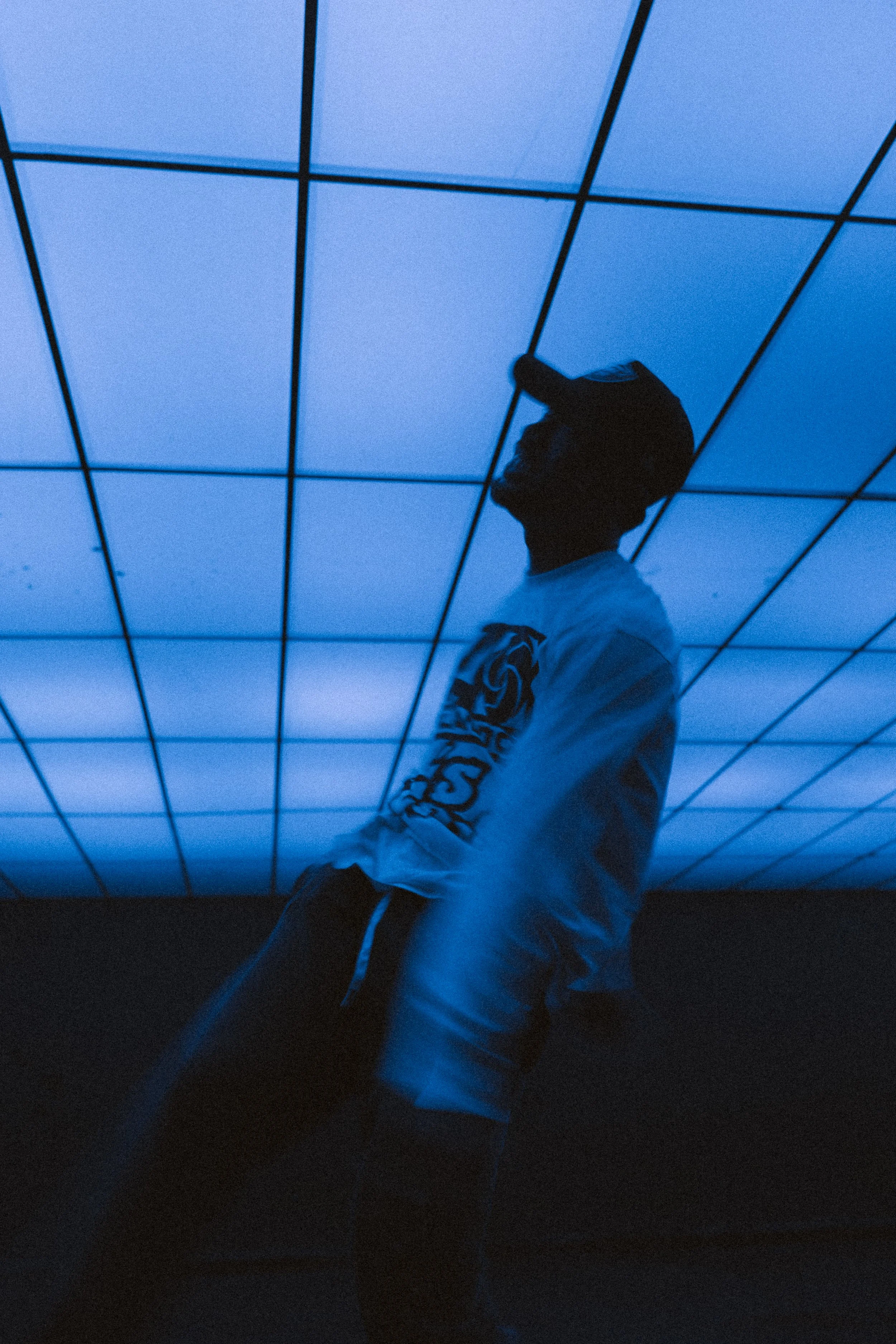 A person leaning against a black surface, wearing a white jacket with a black and white patterned sleeve, a black cap, and black glasses, standing in front of a glowing blue grid ceiling.