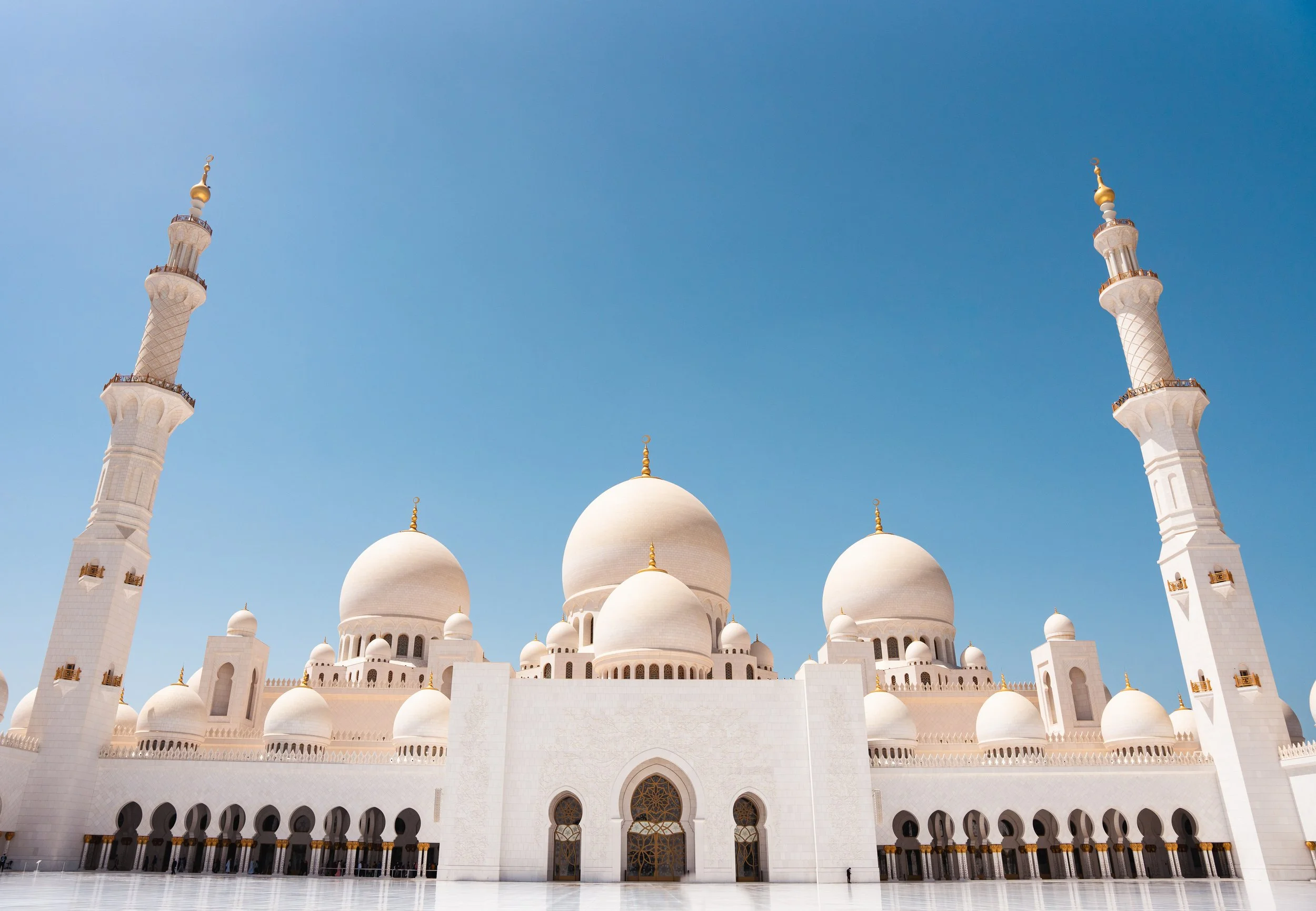 A white mosque with multiple domes and tall minarets against a clear blue sky.