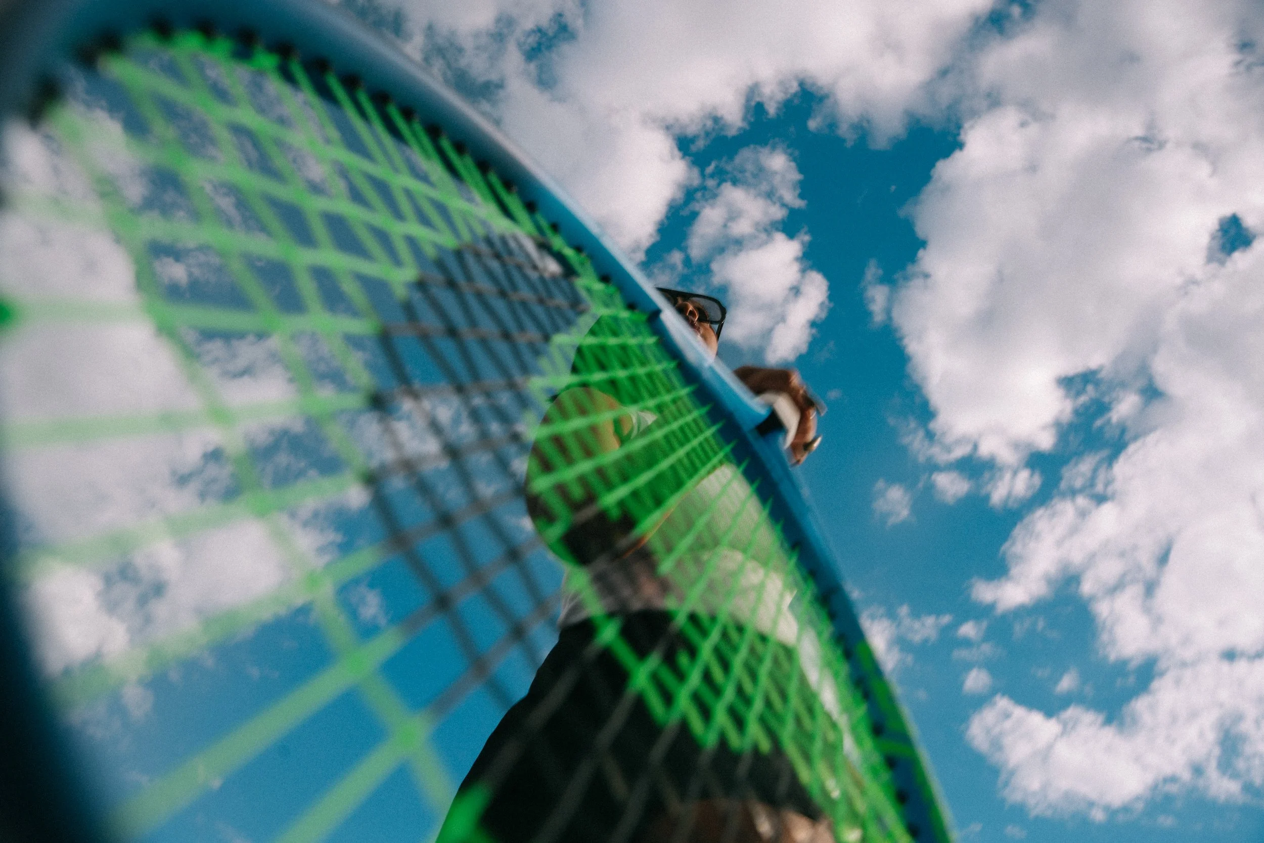 Looking up at a tennis player hitting a ball over a net with blue sky and white clouds in the background.