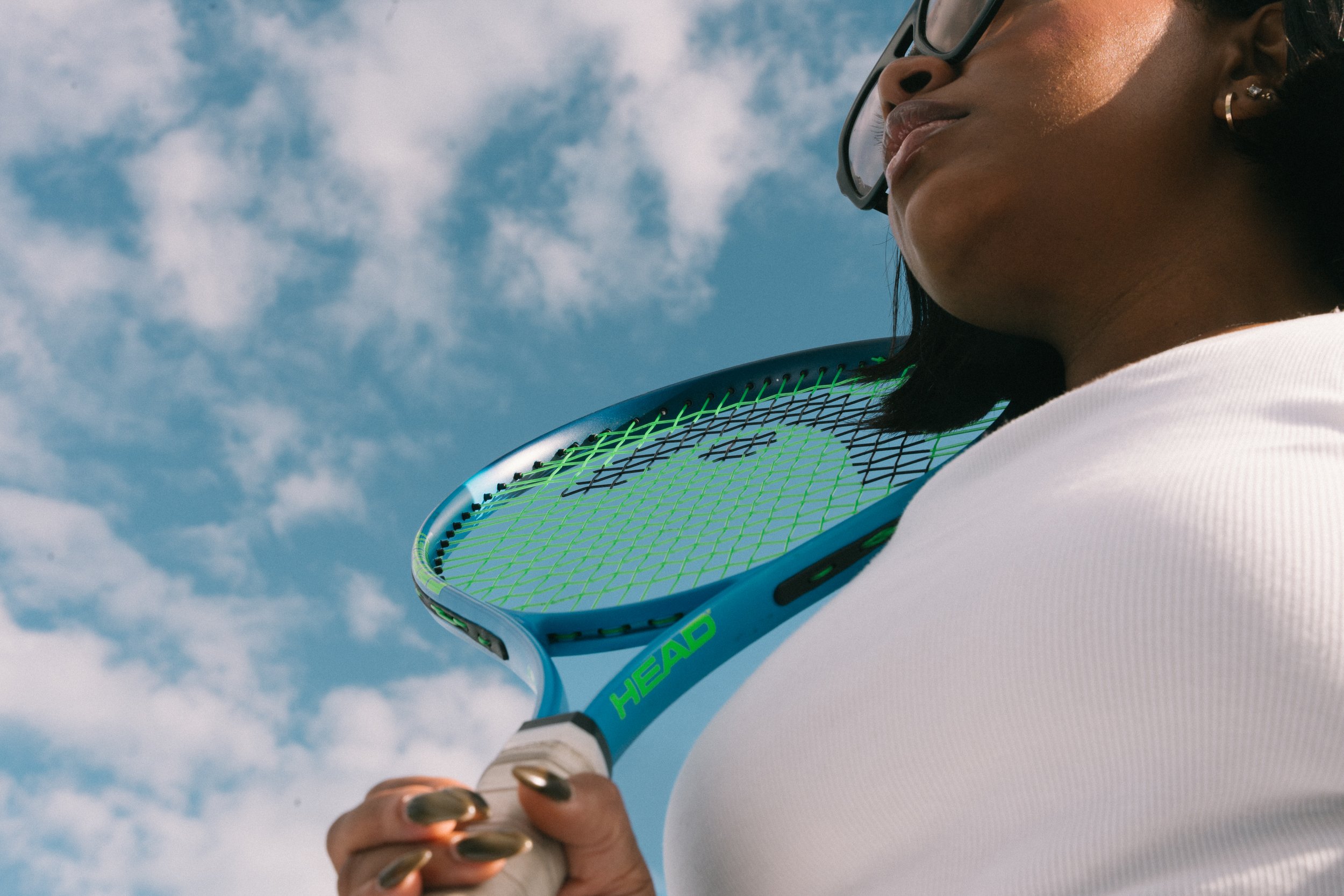A woman wearing glasses and a white shirt holding a tennis racket with a blue frame and green strings against a partly cloudy sky.