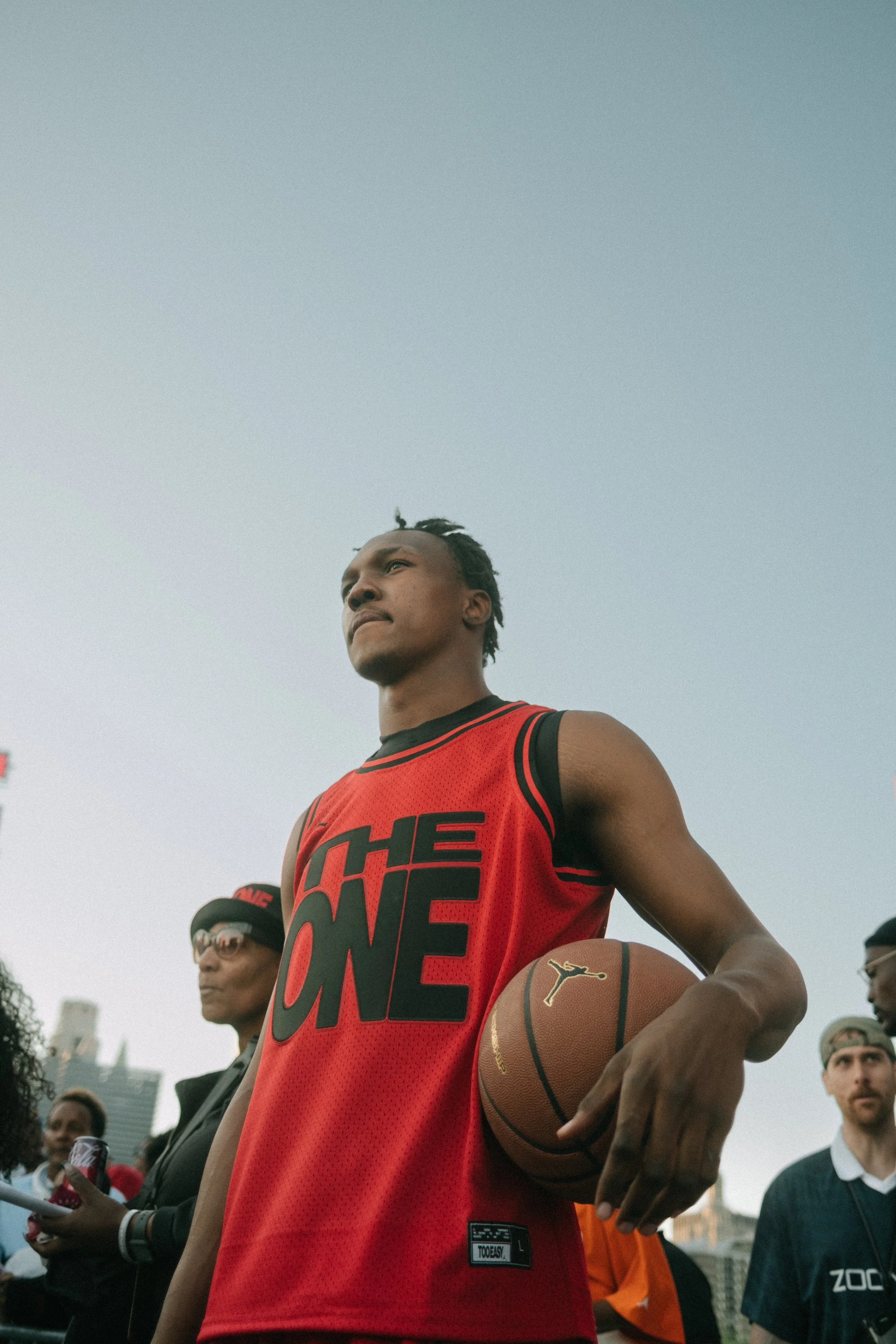 A young man in a red sports jersey holding a basketball, standing outdoors with a crowd and city buildings in the background.