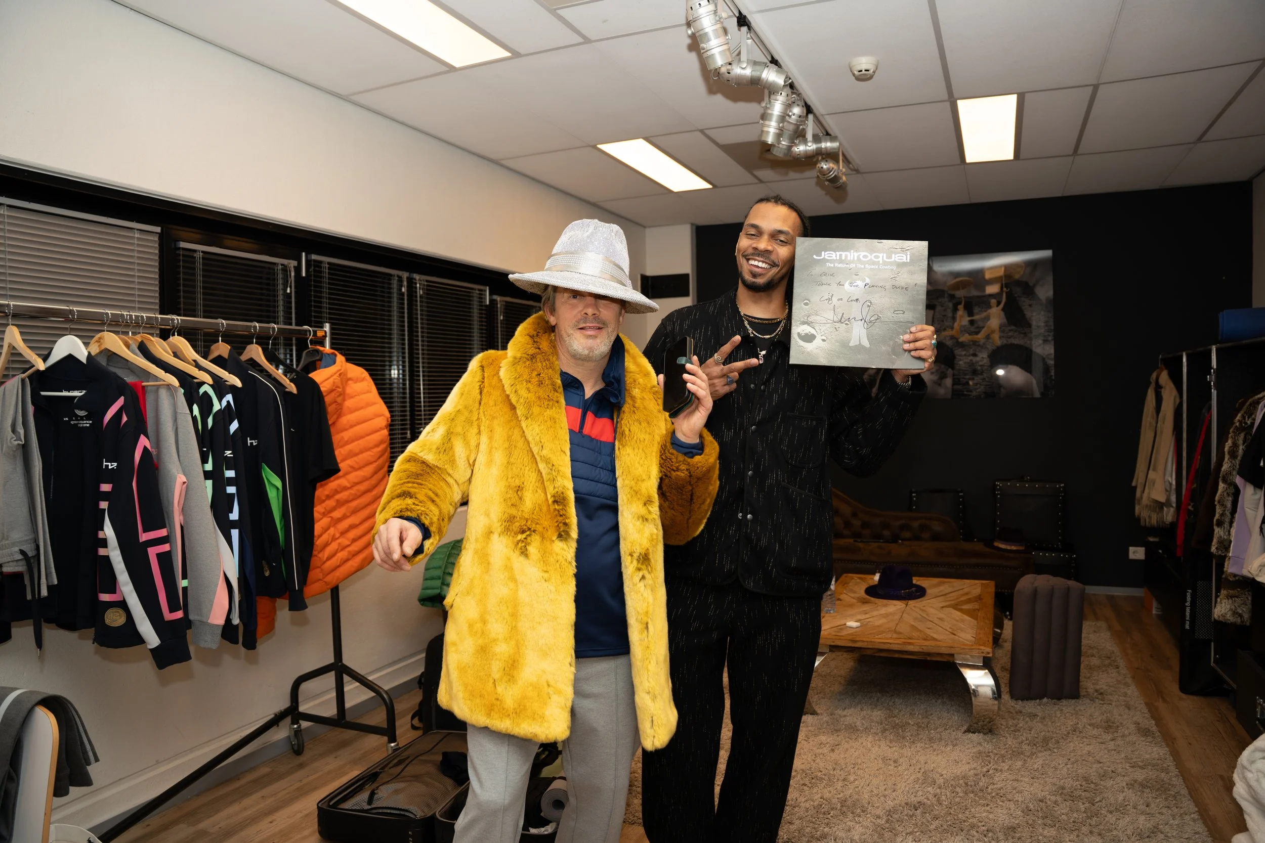 Two men are standing in a room filled with clothing, one wearing a bright yellow fur coat and a silver hat, the other holding a record and making a peace sign. They are smiling and posing for the camera.