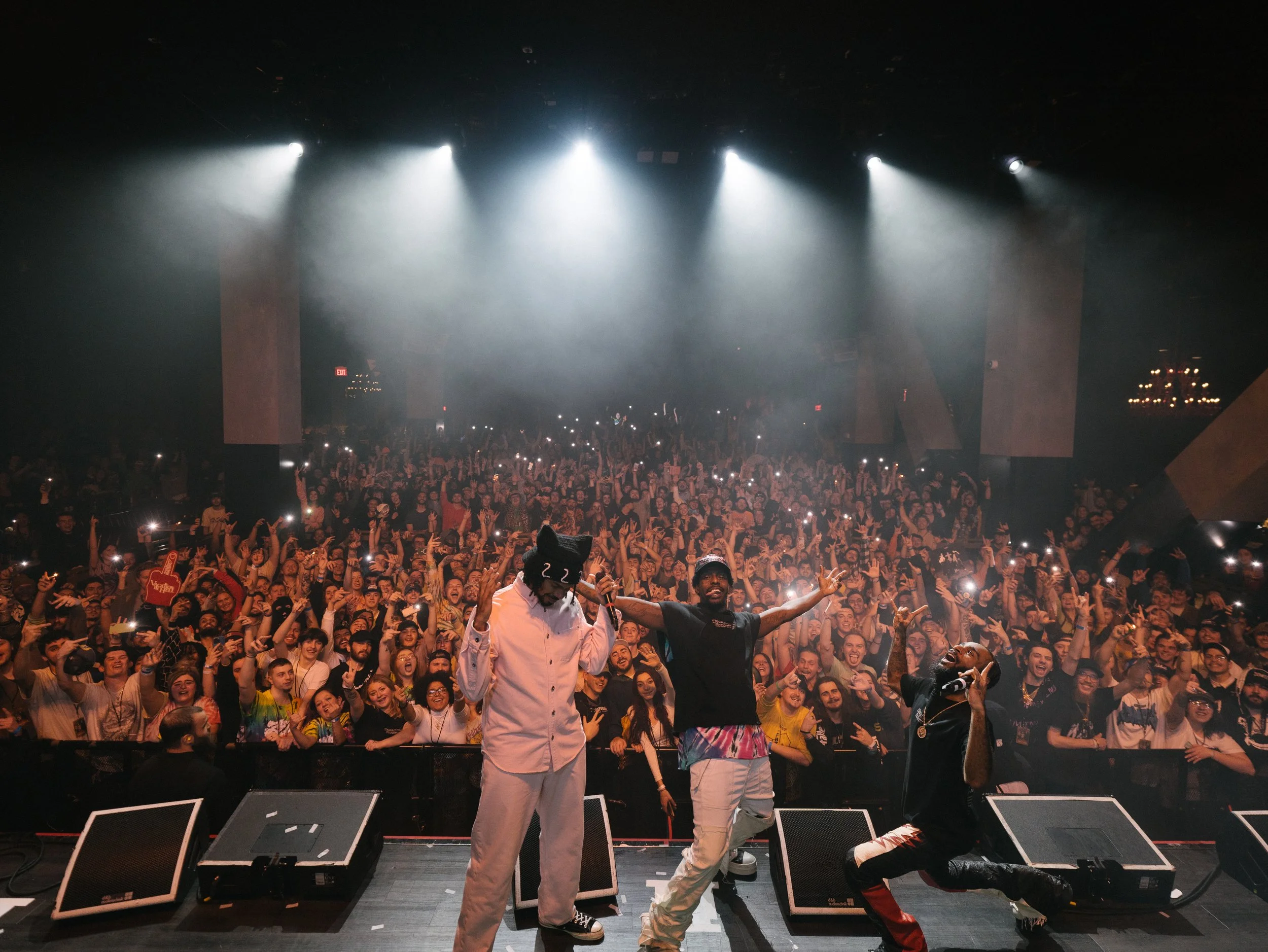 A lively concert scene with three performers on stage performing for a large, cheering audience in a darkened venue.