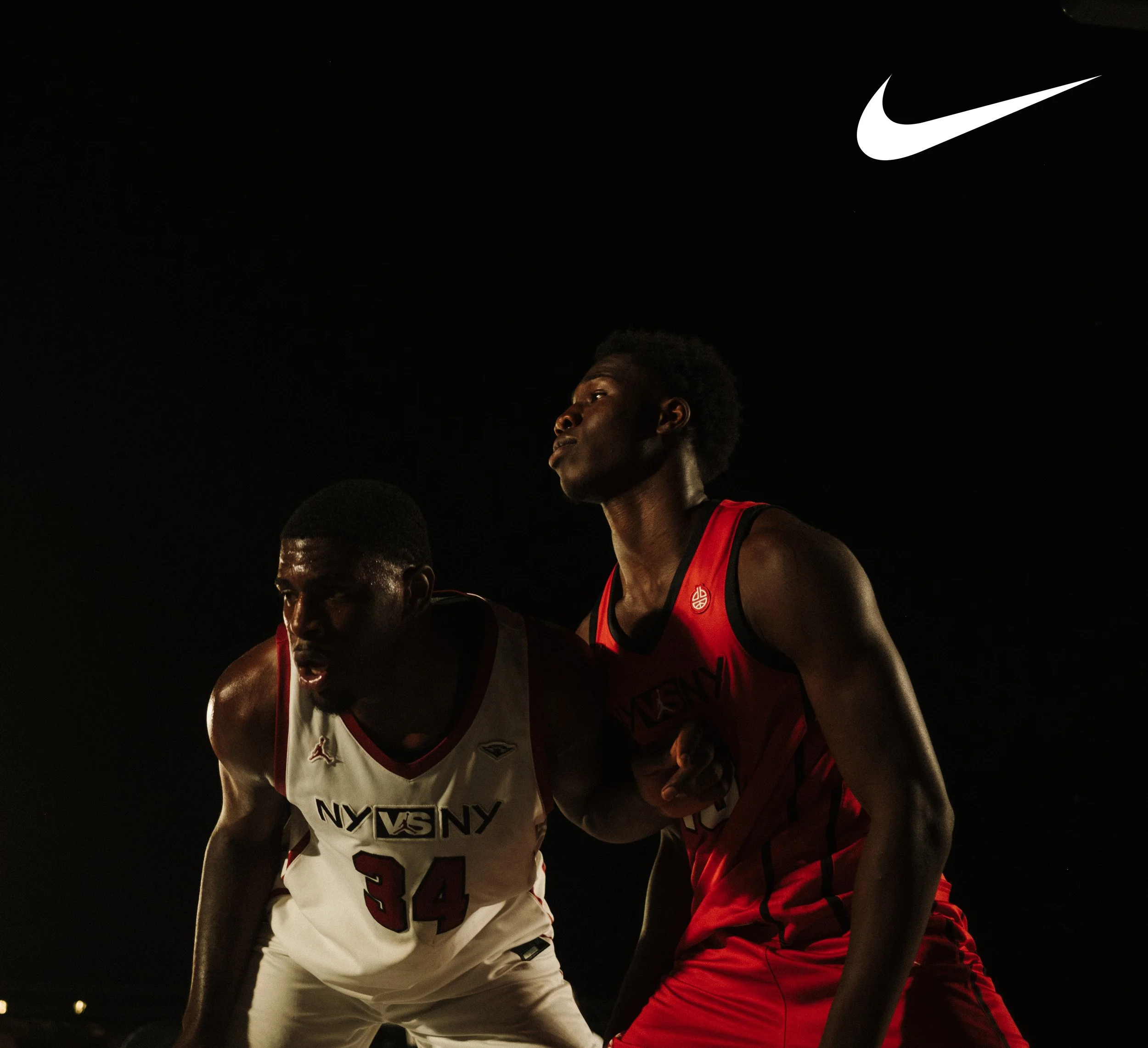 Two male basketball players competing during a game at night, one in a white jersey with the number 34 and the other in a red jersey, with a Nike logo visible in the dark background.
