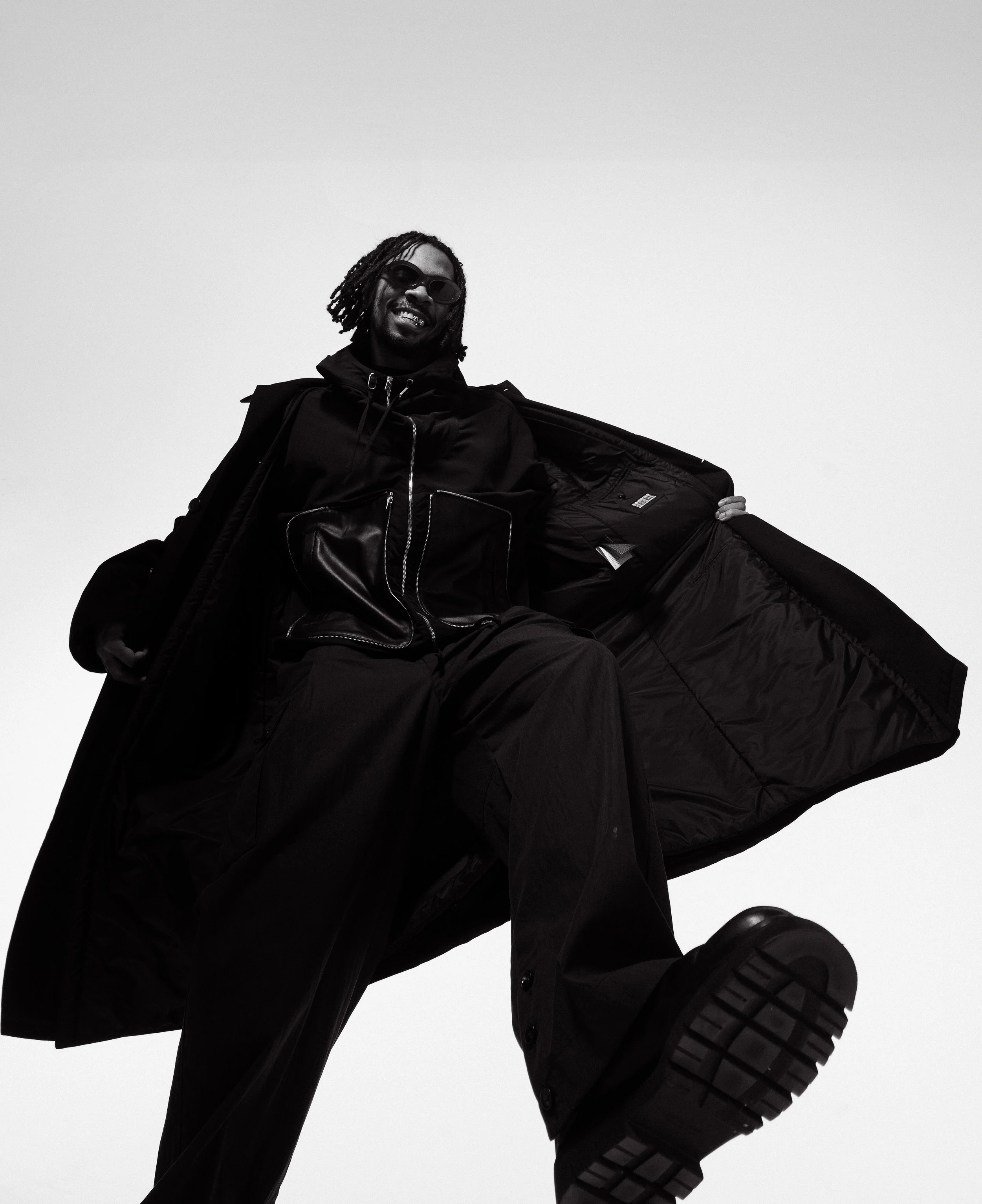 A man with dreadlocks wearing sunglasses, a zip-up jacket, and baggy pants, sitting in a large open black jacket or coat, against a plain background, looking down at the camera with a smile.