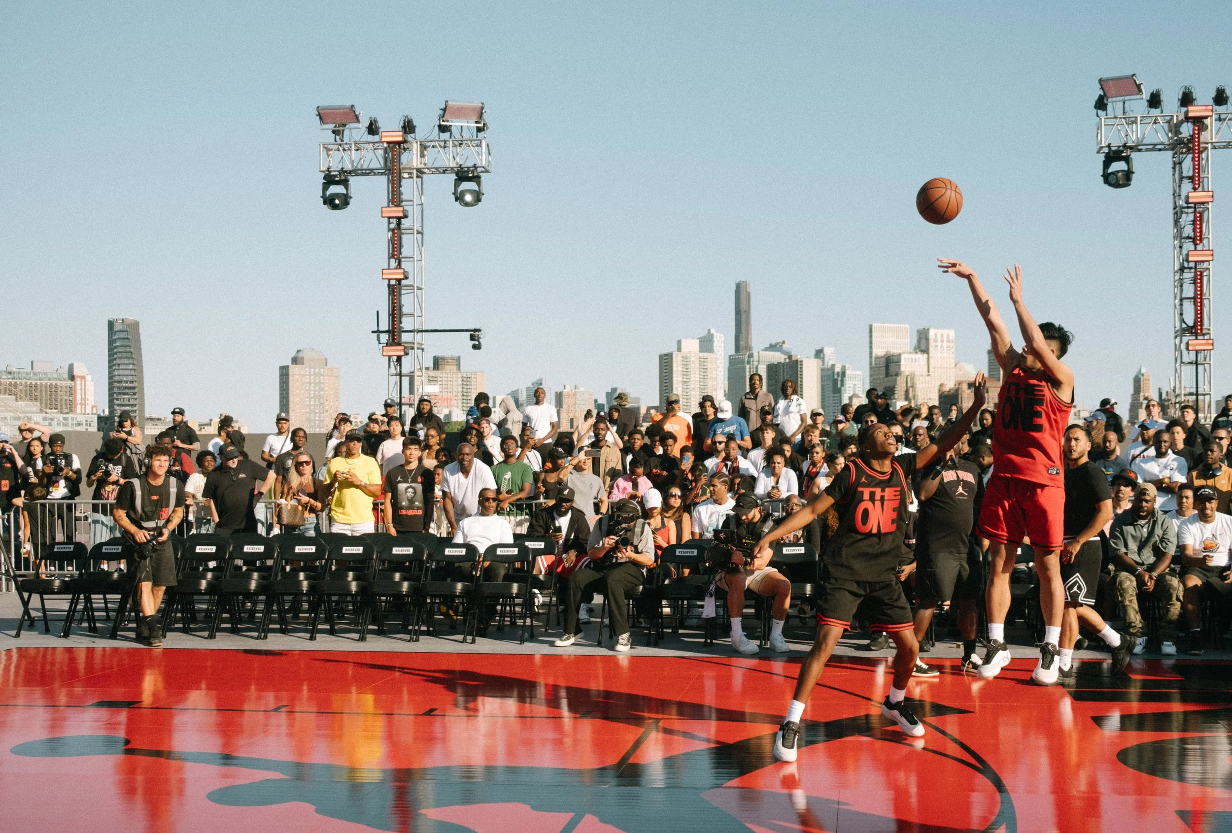 Outdoor basketball game with two players jumping for a shot or block, surrounded by a crowd of spectators and city skyline in the background.