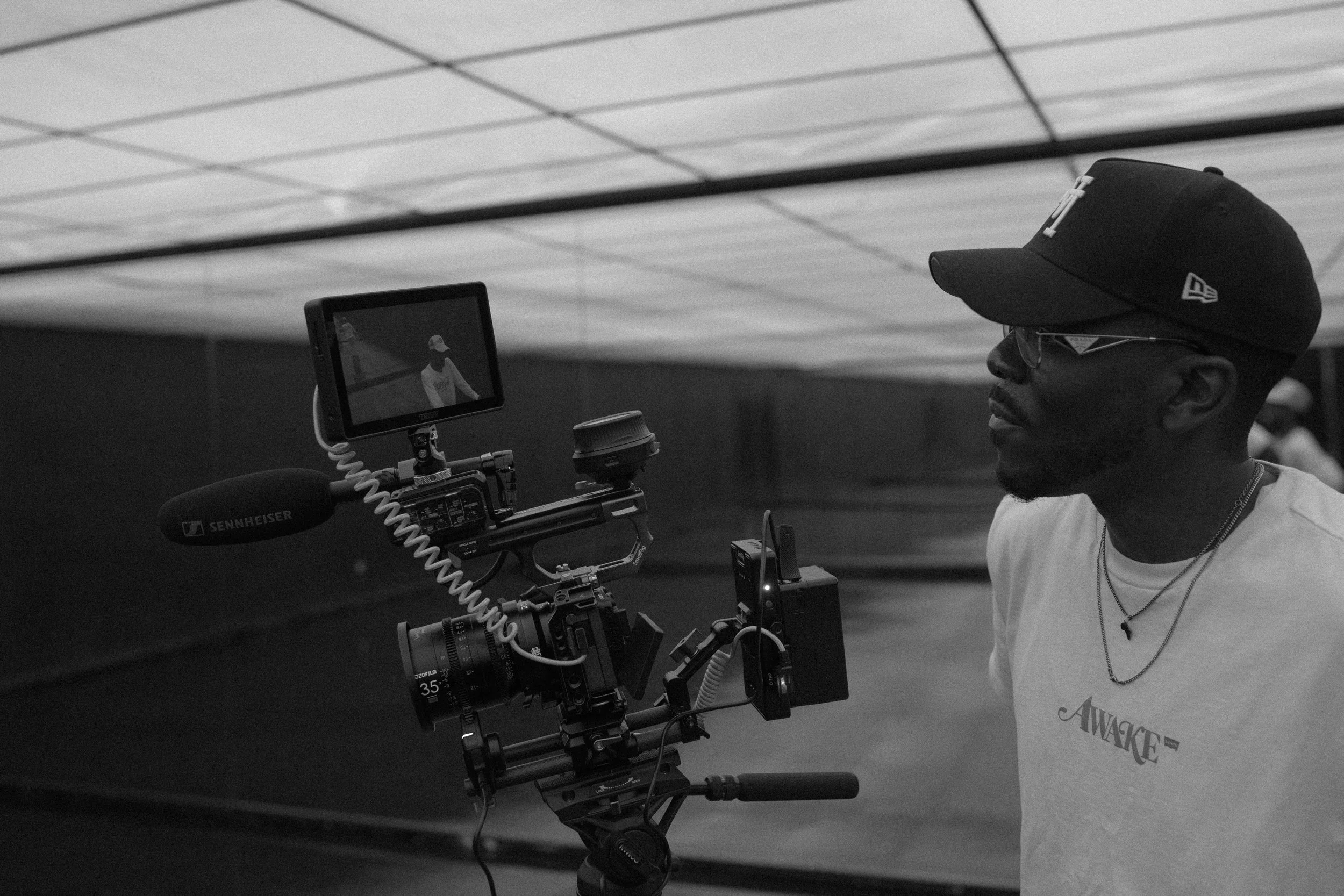 A man wearing glasses and a cap stands next to a professional video camera on a tripod in an indoor setting.