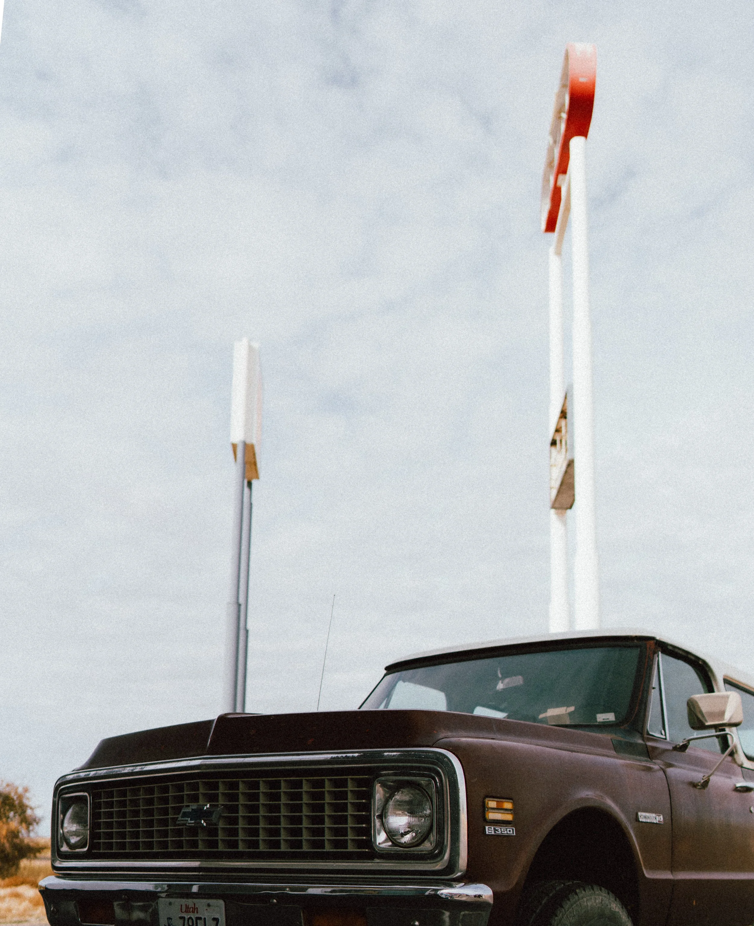 A vintage brown pickup truck parked outdoors near a fast food restaurant, with empty signs on tall poles in the background and a cloudy sky overhead.