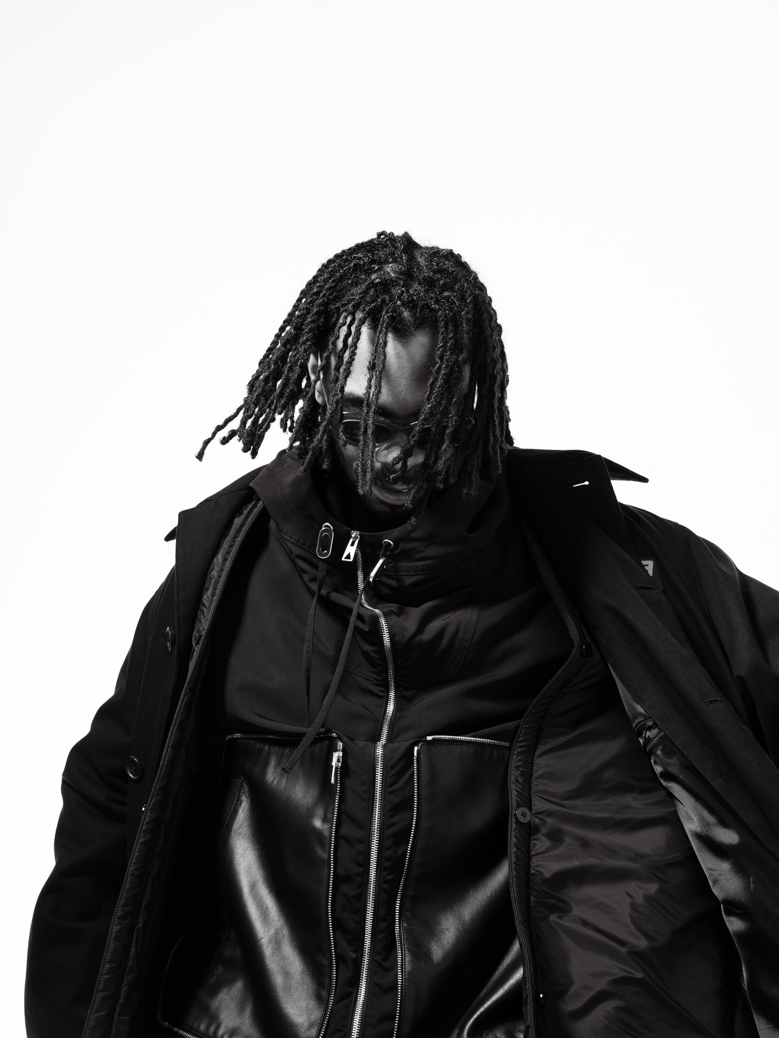 A person with dreadlocks wearing sunglasses and layered black jackets leaning slightly forward against a white background.