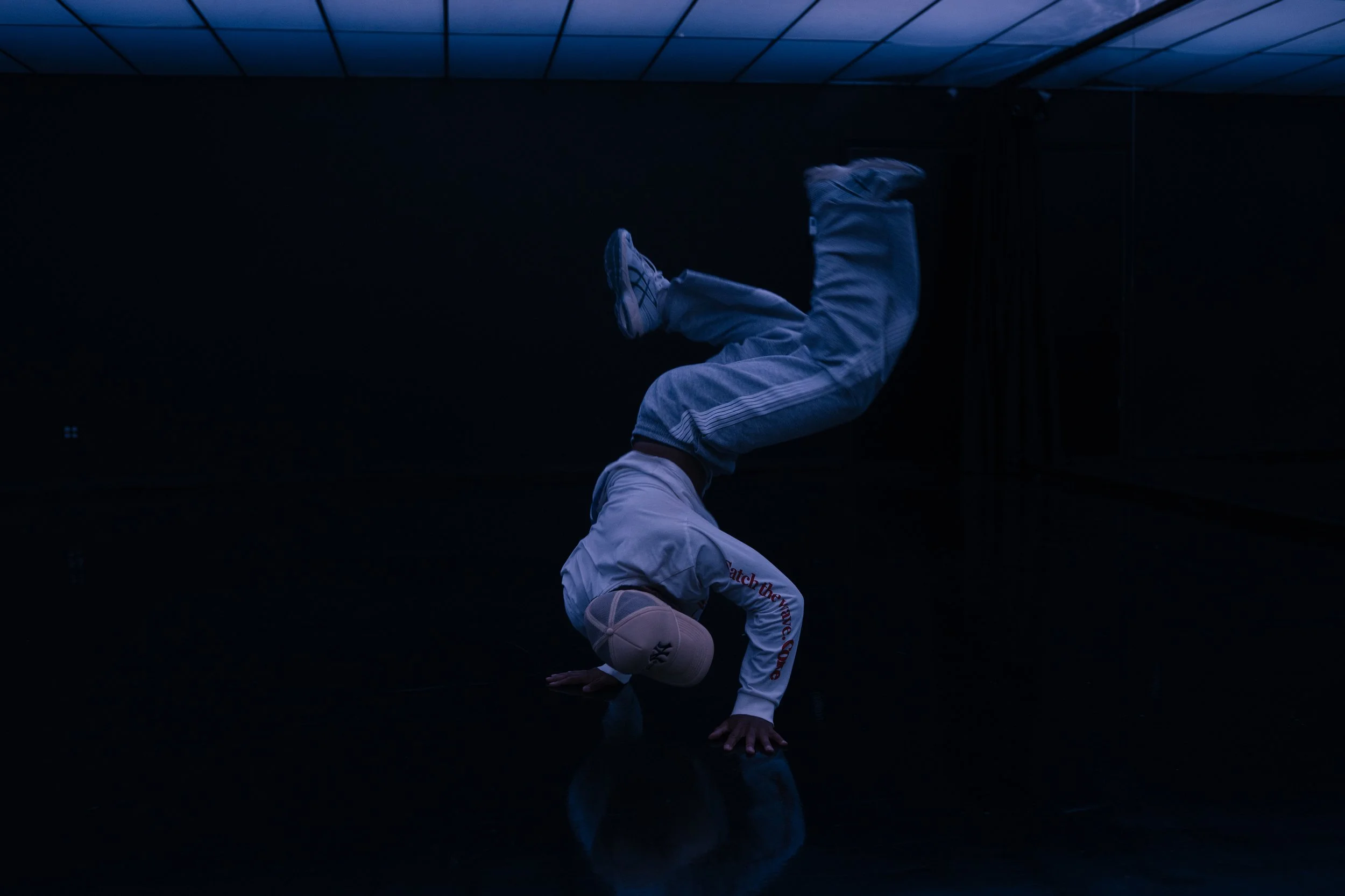 A person dressed in white tracksuit and a beige cap performing a handstand or headstand on a dark, reflective floor in a dimly lit room, possibly a dance or martial arts studio.
