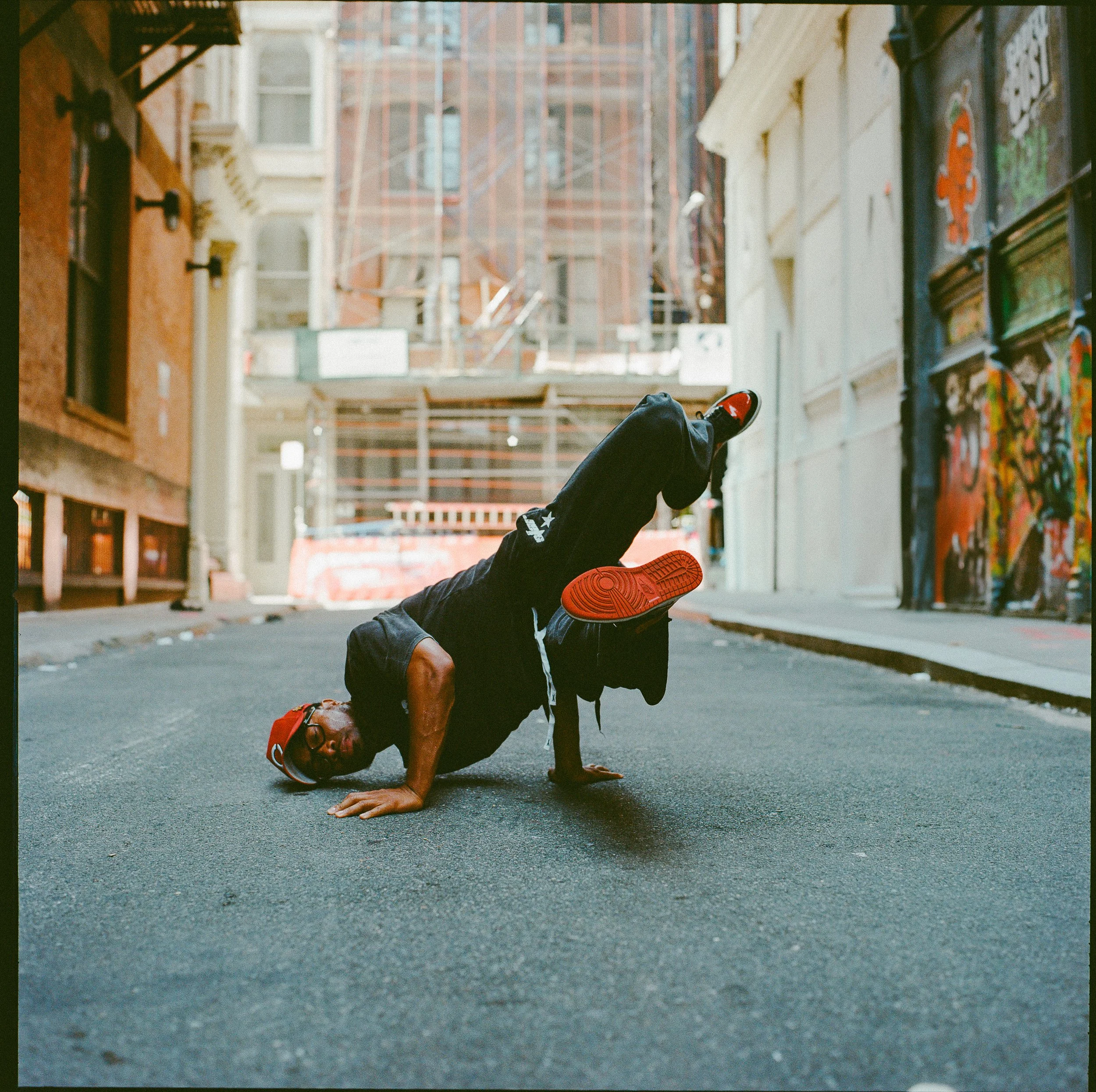 A person performing a handstand in an alleyway with colorful graffiti walls, wearing a black hoodie, black sweatpants, red sneakers, and a red cap.