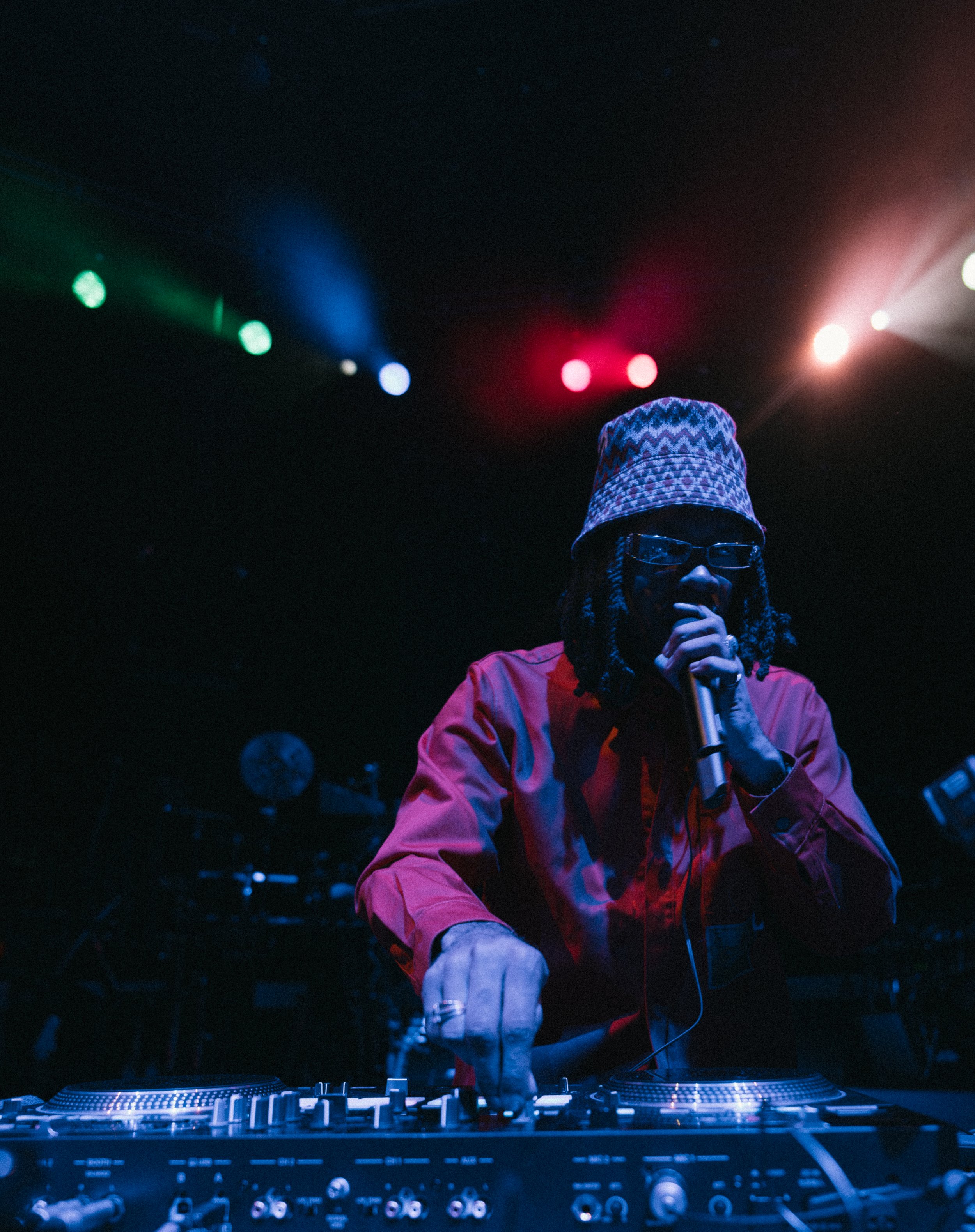 A DJ performing on stage with colorful lights shining above, wearing glasses, a patterned bucket hat, and a red jacket, using DJ equipment.
