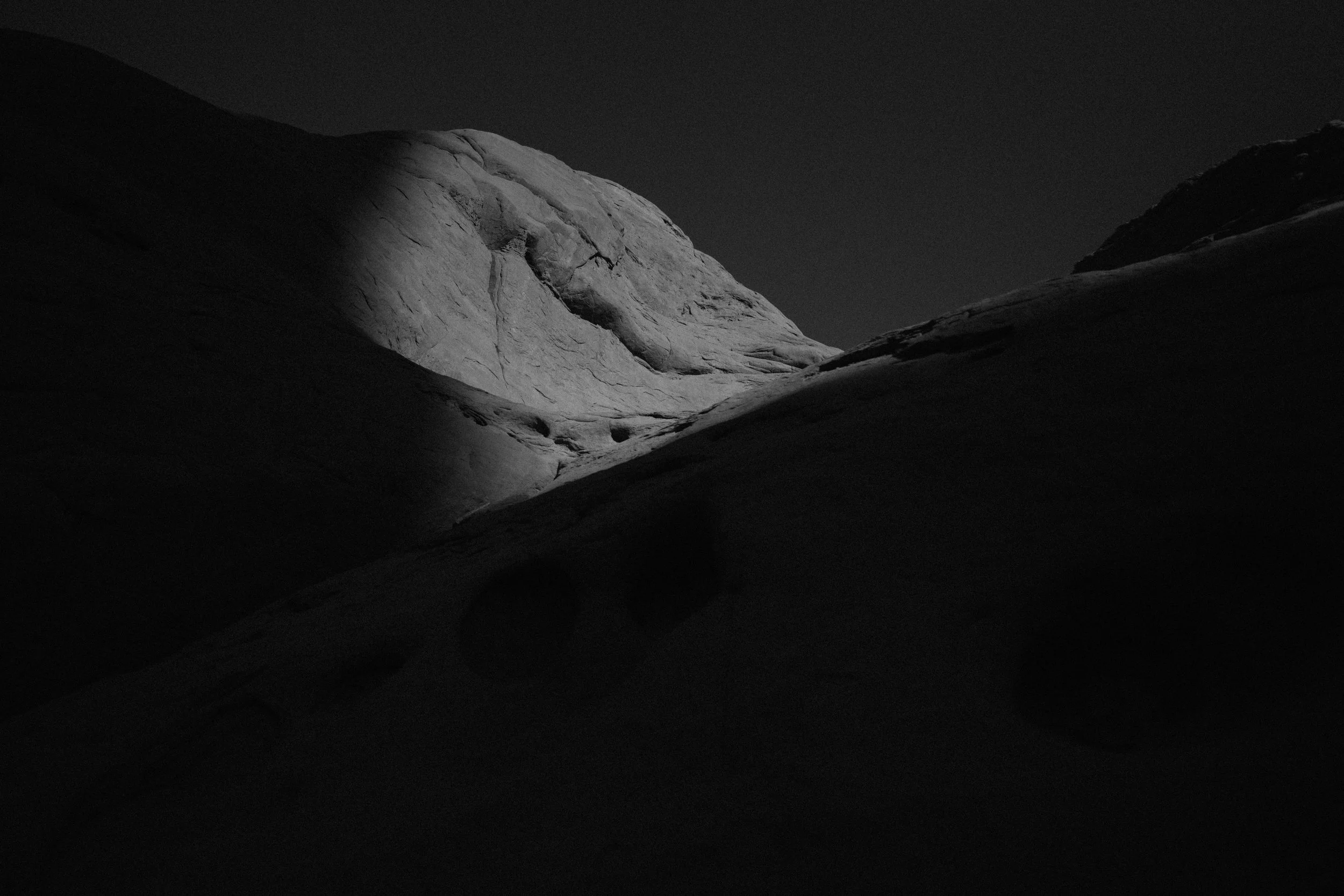 Nighttime photo of a large rocky mountain with a bright illuminated section, dark shadowed slopes in the foreground, and a dark sky.