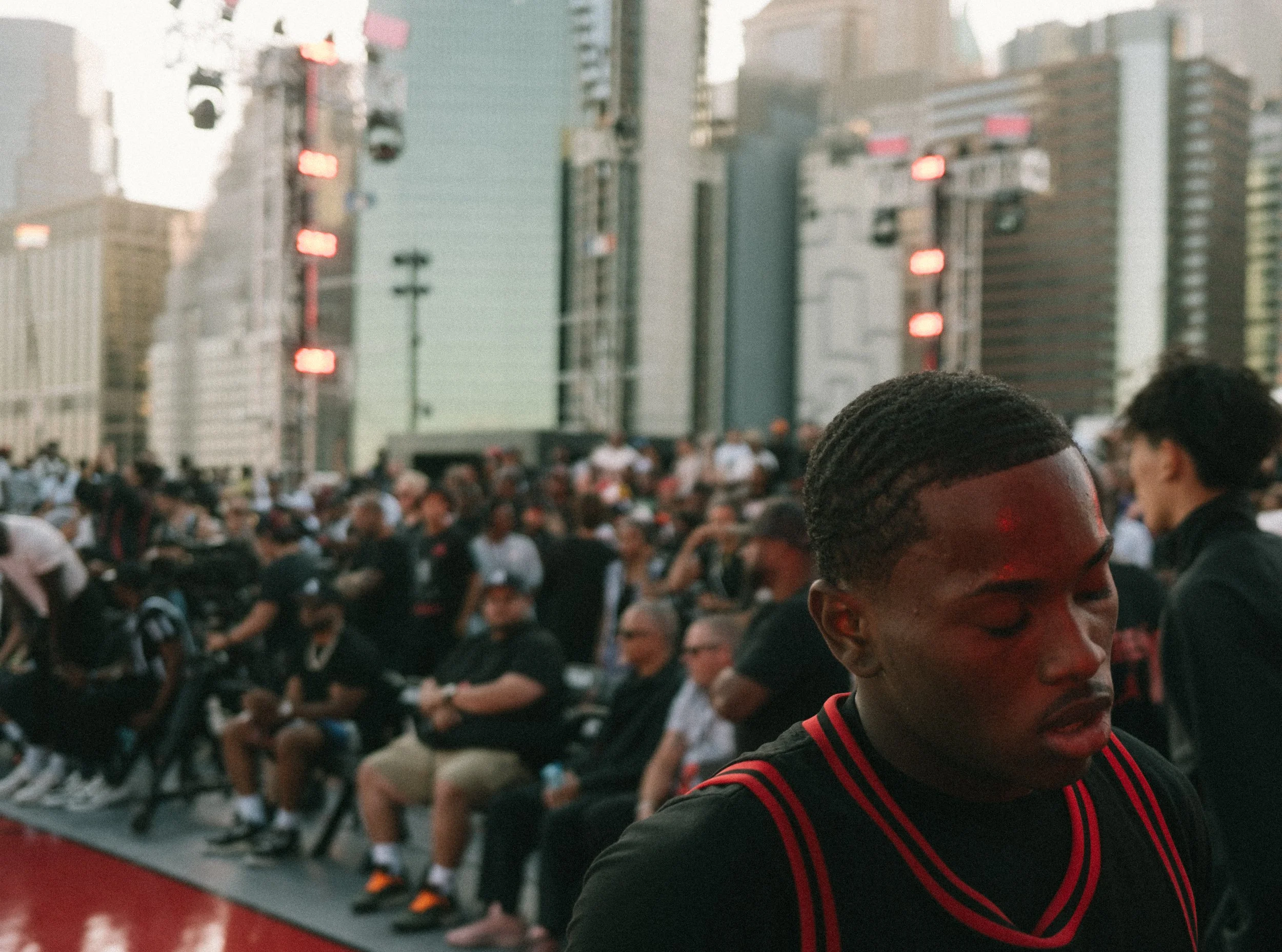 A man with braided hair wearing a black sports shirt with red stripes, standing in front of a crowd at an outdoor basketball game, with tall buildings in the background.