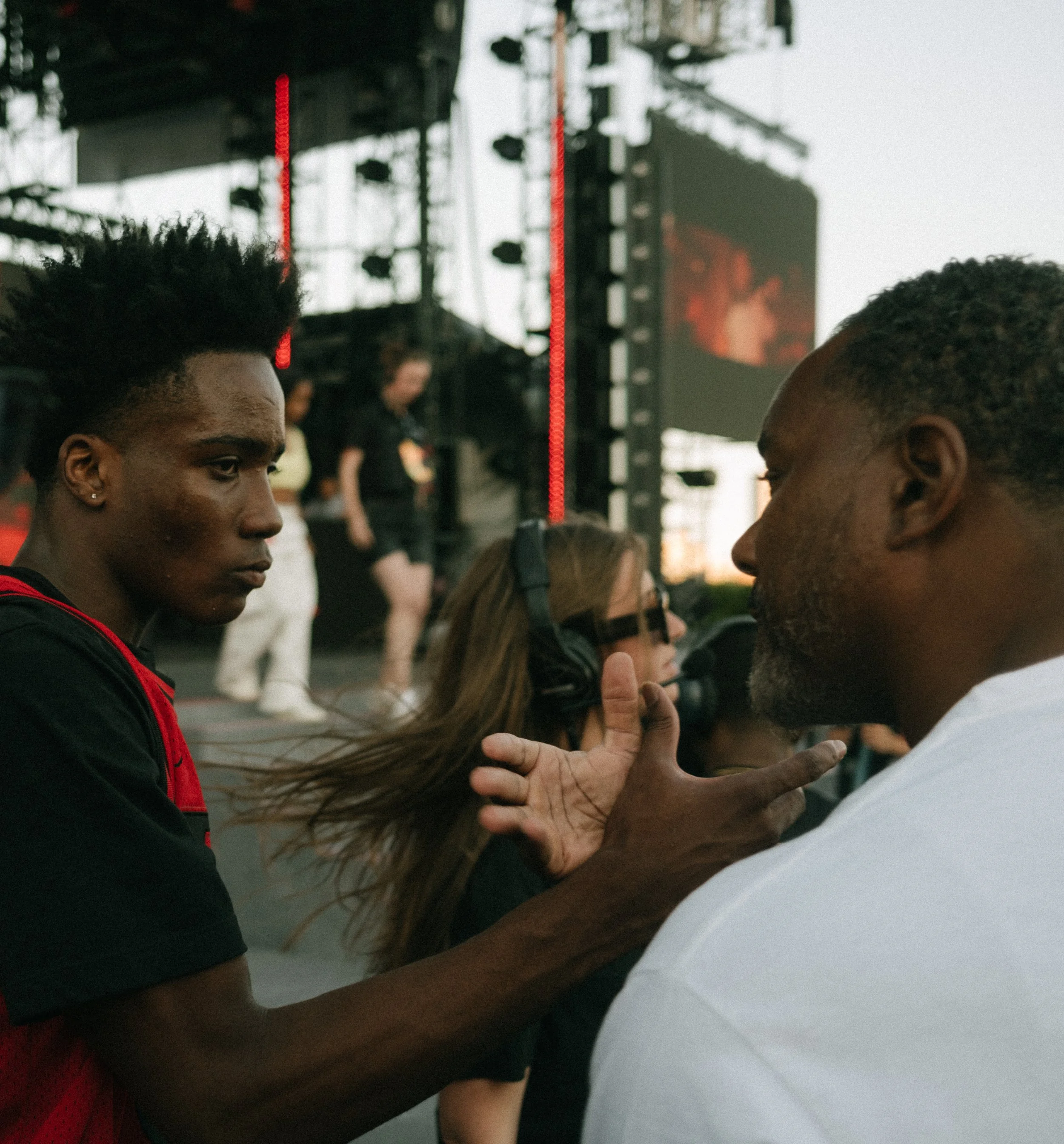 Two men are engaged in a serious conversation at an event. One man has dark skin, black hair, and wears a black and red shirt. The other man has slightly lighter skin, short hair, and a beard, wearing a white shirt. In the background, a woman with lo