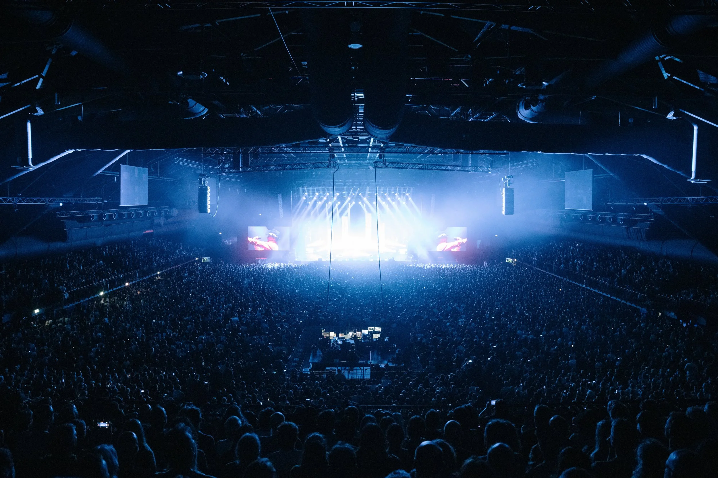 Large concert crowd inside an indoor arena with a bright stage and screens.