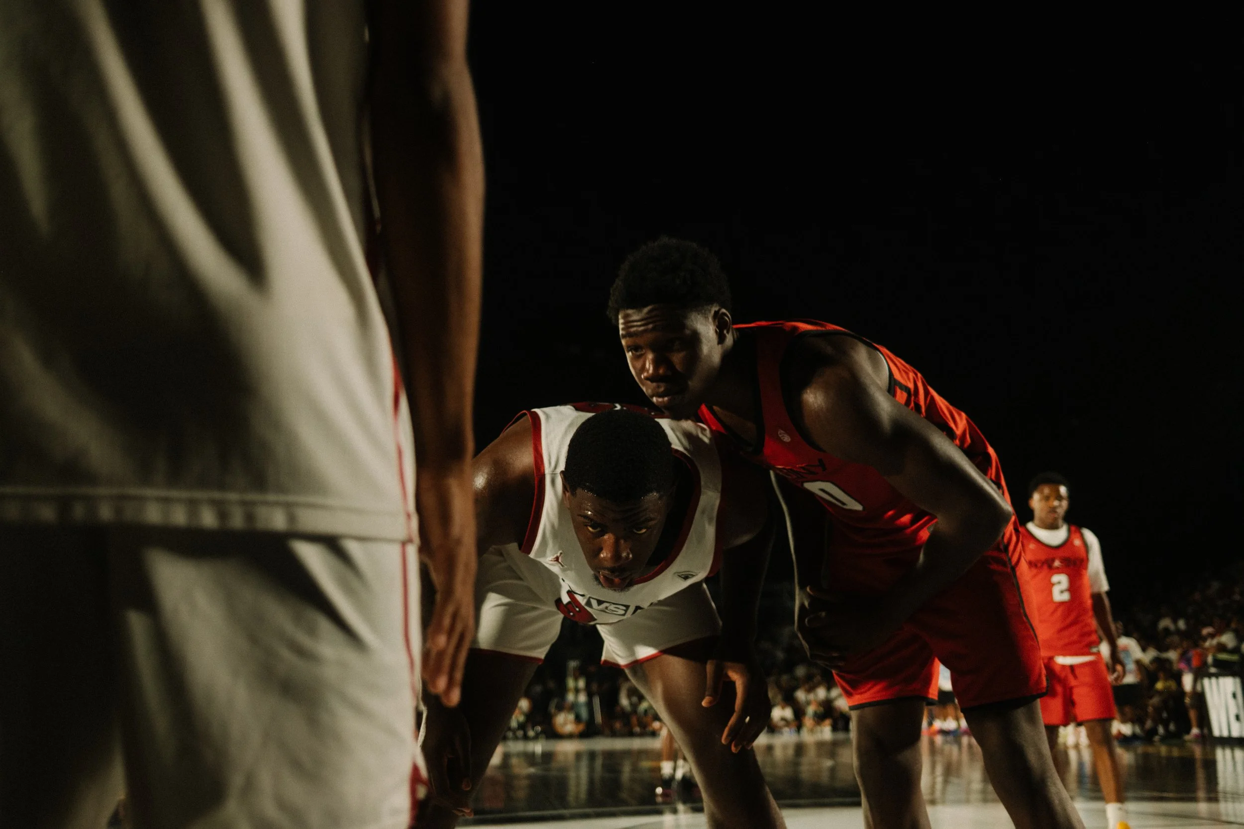 Two basketball players, one in a white jersey, bending forward and looking intense, and the other in a red jersey, with a determined expression, face off on a basketball court at night, with yellow-jerseyed player visible in the background.