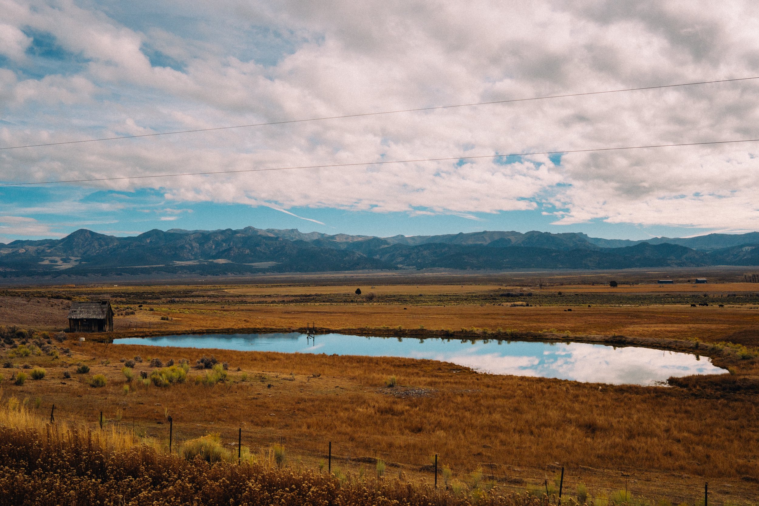 A vast rural landscape with a small pond reflecting the sky, a weathered barn on the left, open fields, and mountains in the distance under partly cloudy skies.