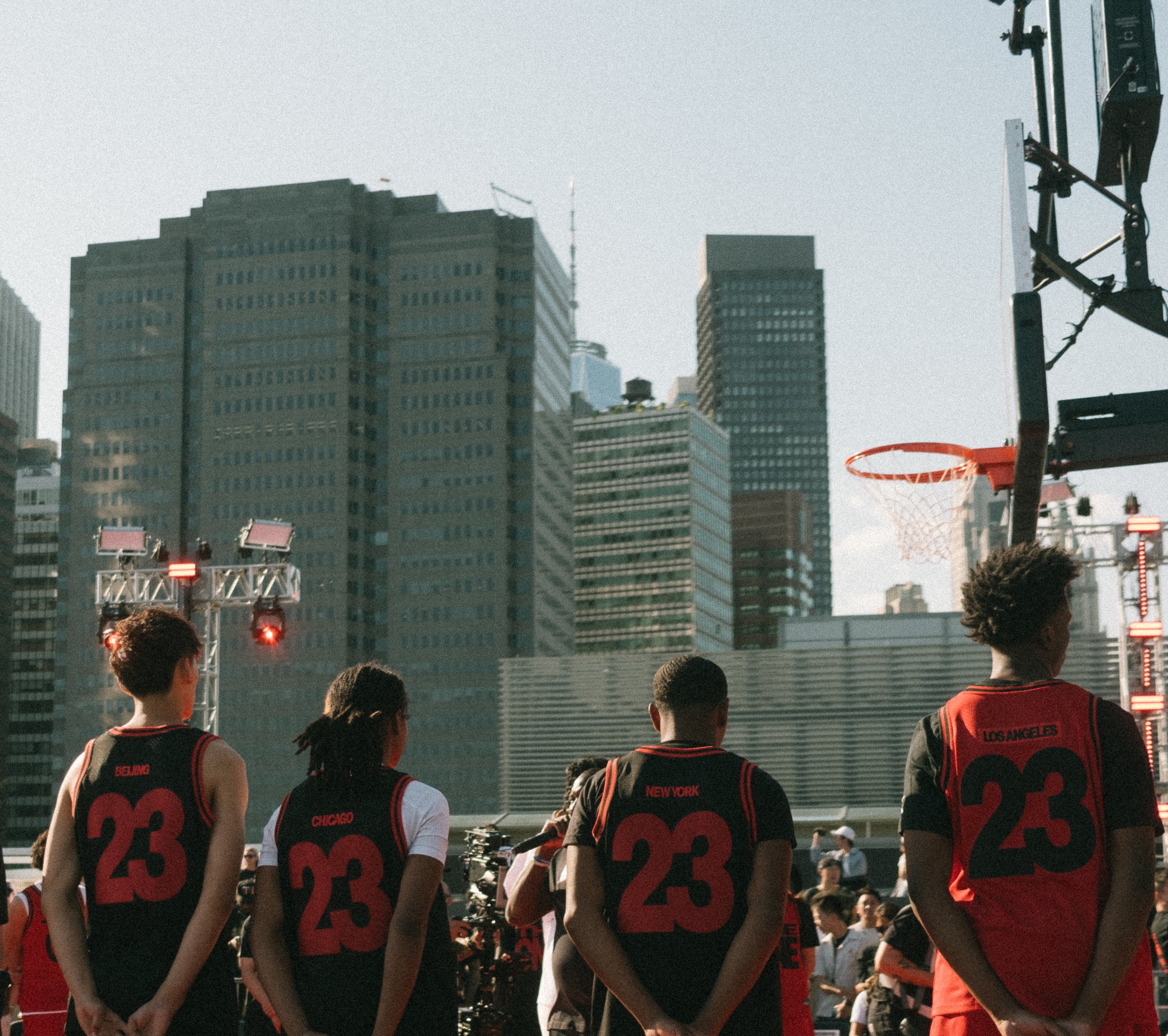 Four young basketball players standing on an outdoor court with city skyscrapers in the background, wearing black and red uniforms with the number 23 and city names.