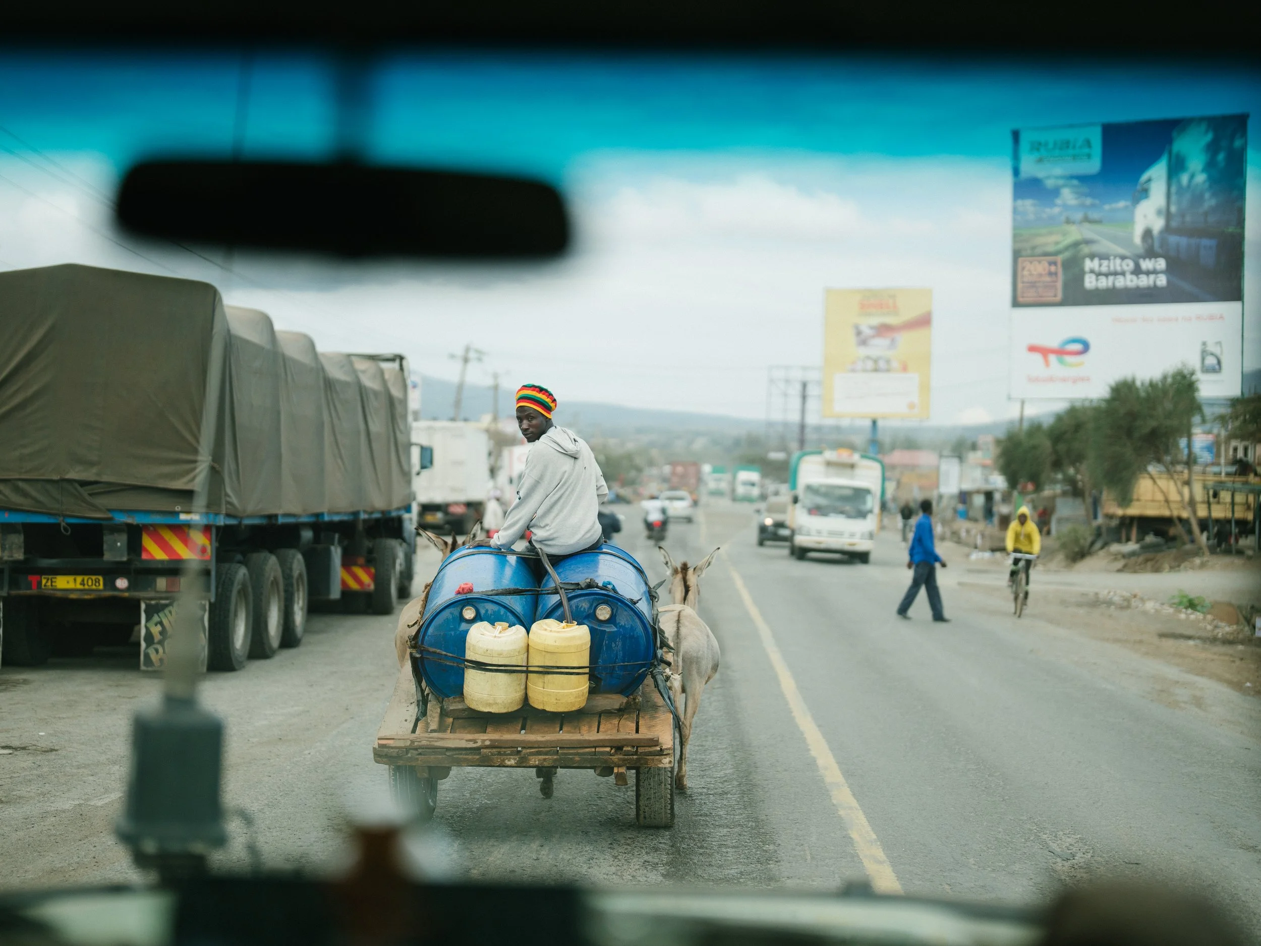 A man riding a donkey cart on a busy road, carrying blue water tanks and yellow containers, with trucks and pedestrians visible in the background.