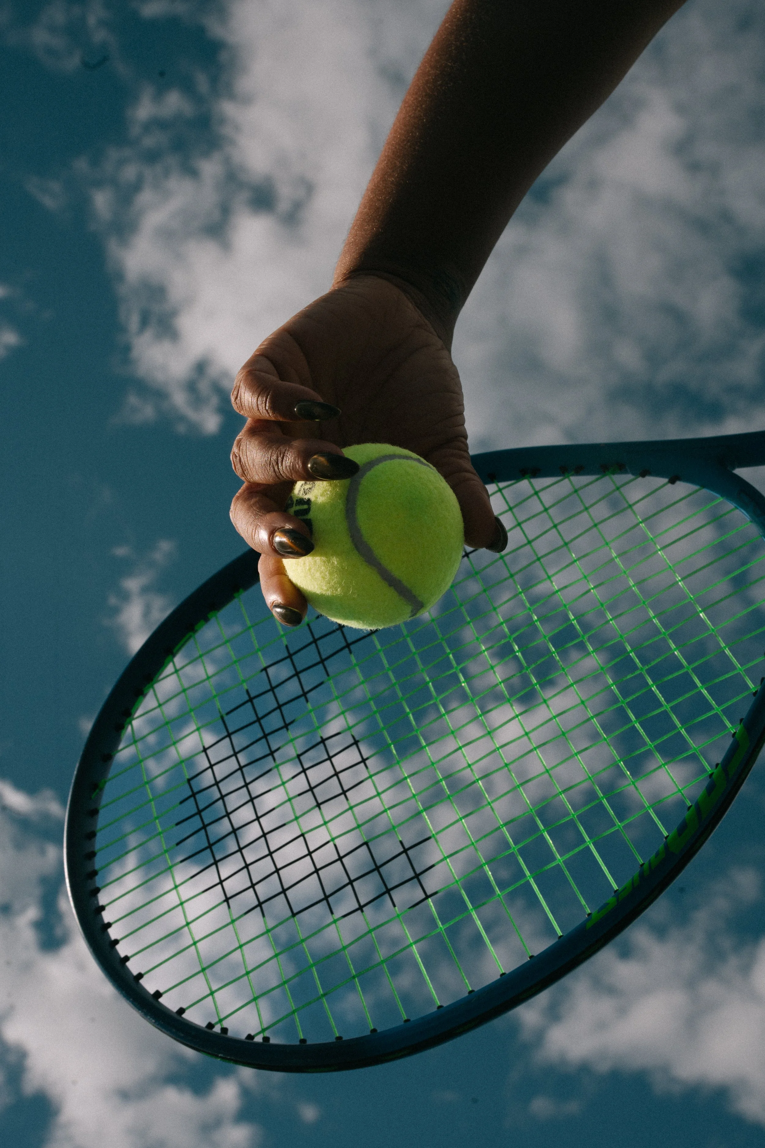 Close-up of a person holding a tennis ball and a tennis racket against a cloudy sky, photographed from below.