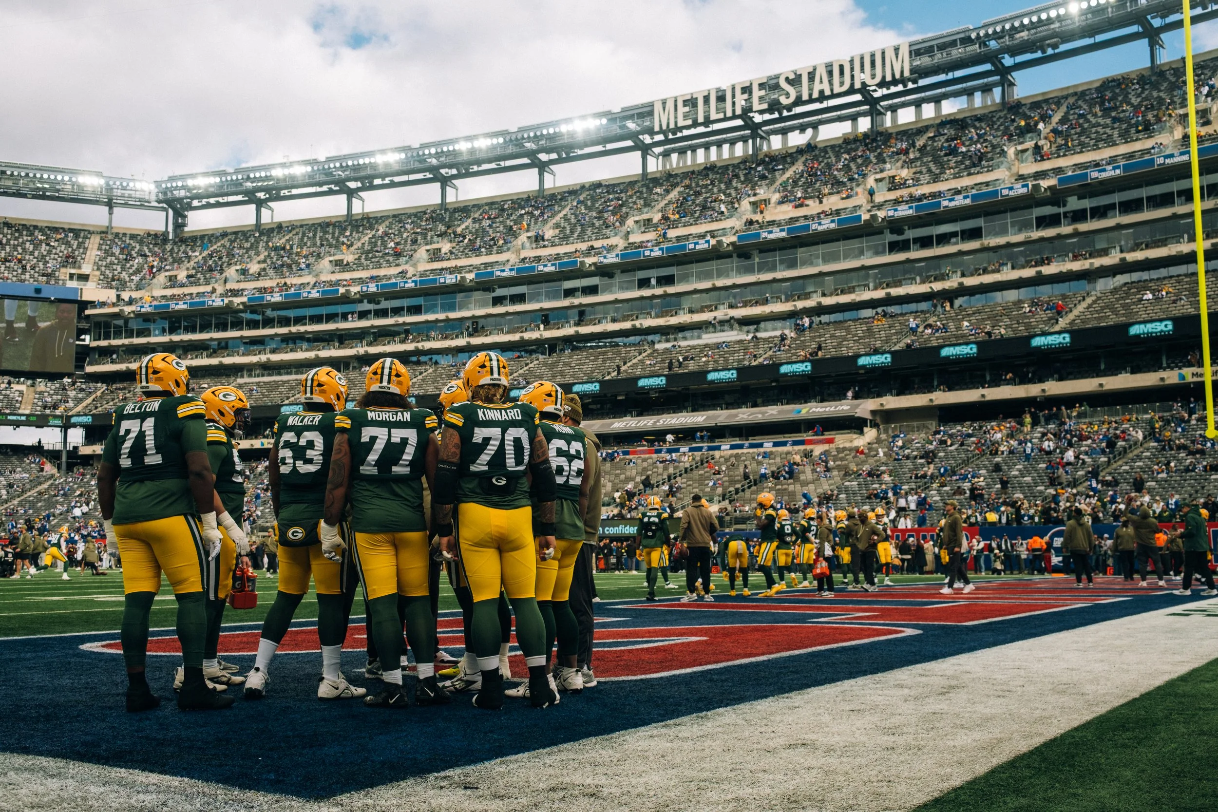 Green Bay Packers football players standing on the sideline during a game at MetLife Stadium with a crowd in the stands.