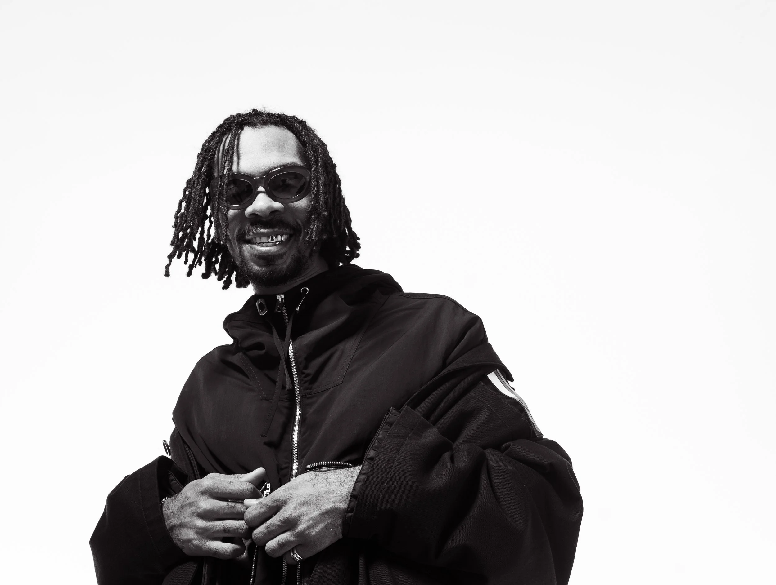 Black and white photo of a smiling man with dreadlocks wearing sunglasses and a black jacket.