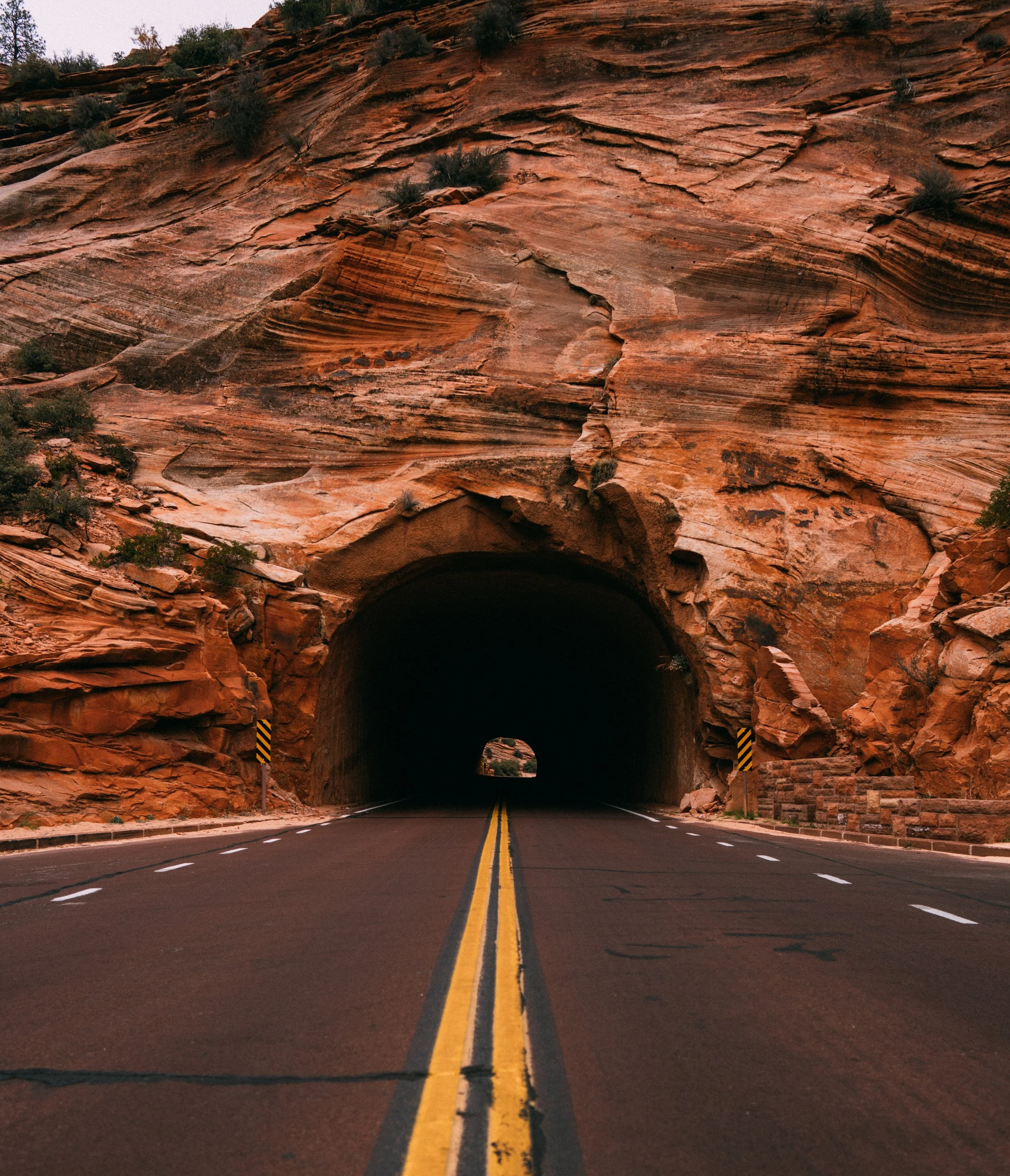 A two-lane road with yellow dividing lines runs through a tunnel carved into red rock formations in a desert landscape.