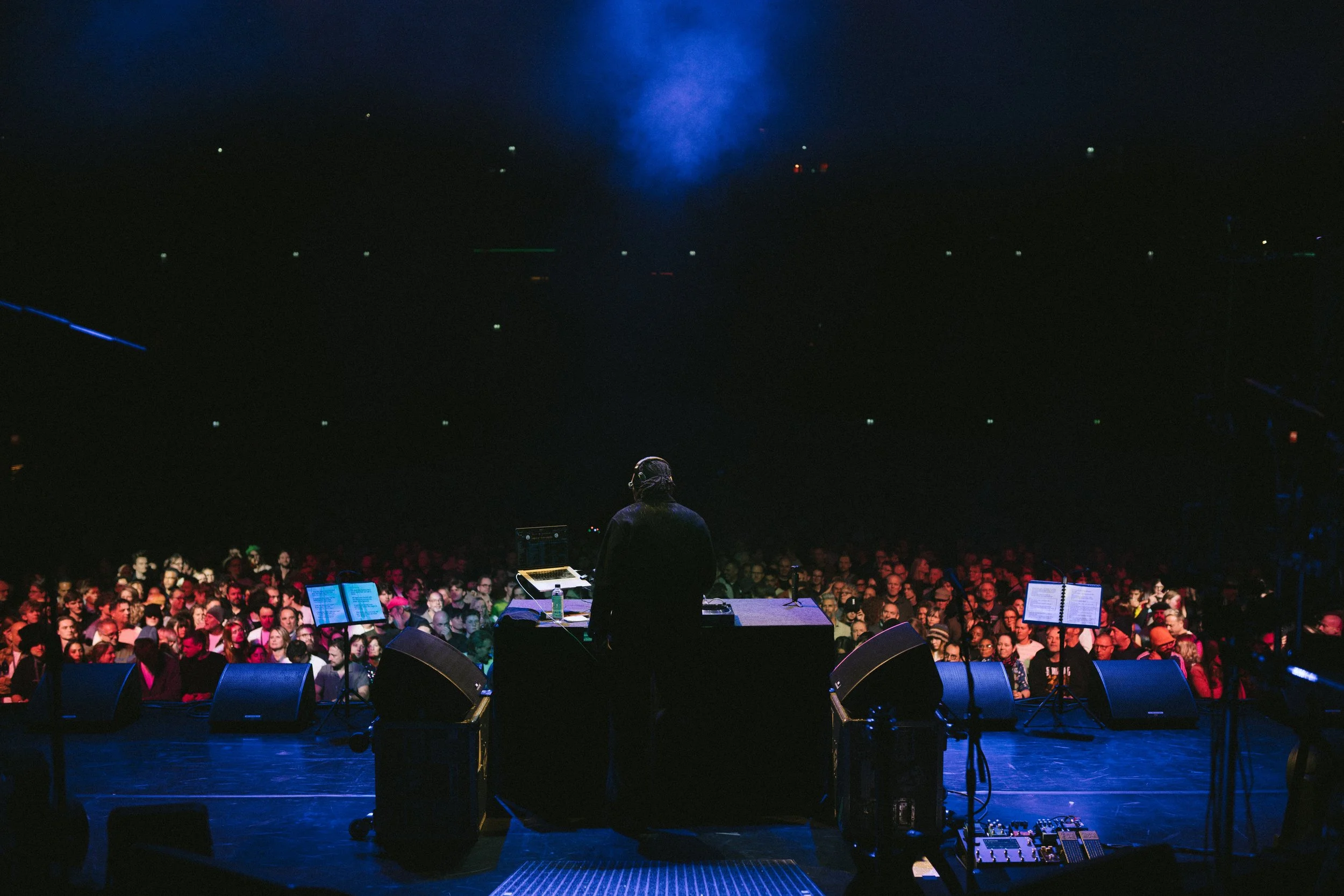 A performer on stage in front of a large audience at a concert or event, with stage equipment and music stands visible.