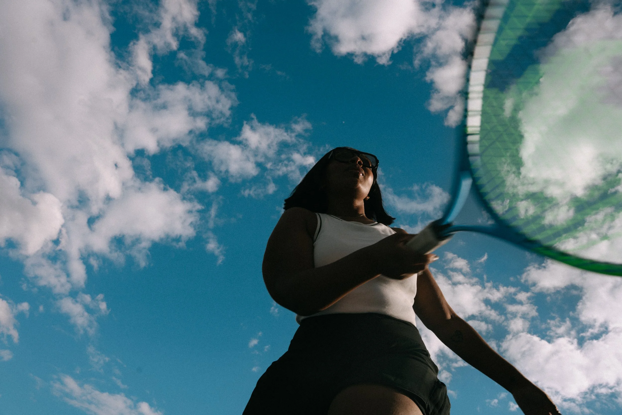 Low angle view of a woman holding a tennis racket, facing the sky with scattered clouds.