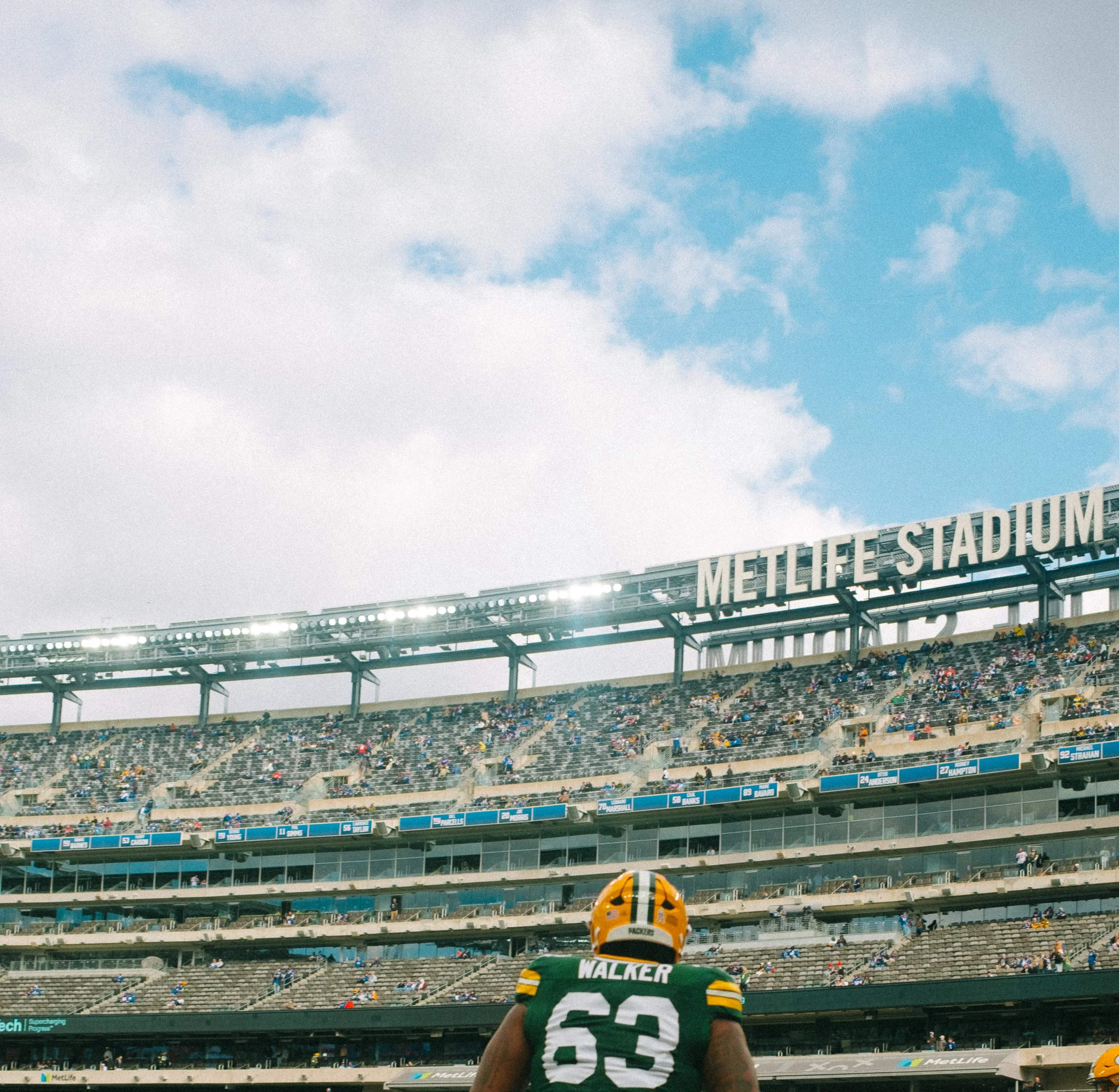 A football player wearing a green and yellow uniform with the name 'Walker' and number 63 on the back, standing on the field at MetLife Stadium with empty and crowded seats, and a large 