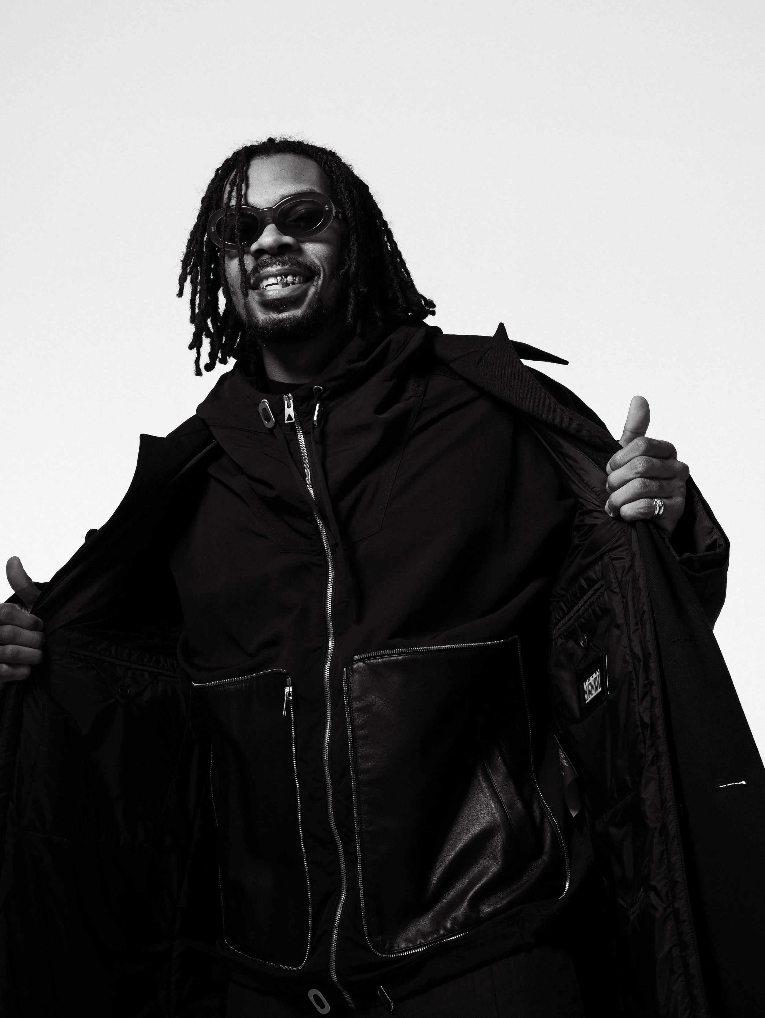 Black and white photo of a man with dreadlocks wearing sunglasses, a black jacket, and showing a confident smile, holding open his jacket.