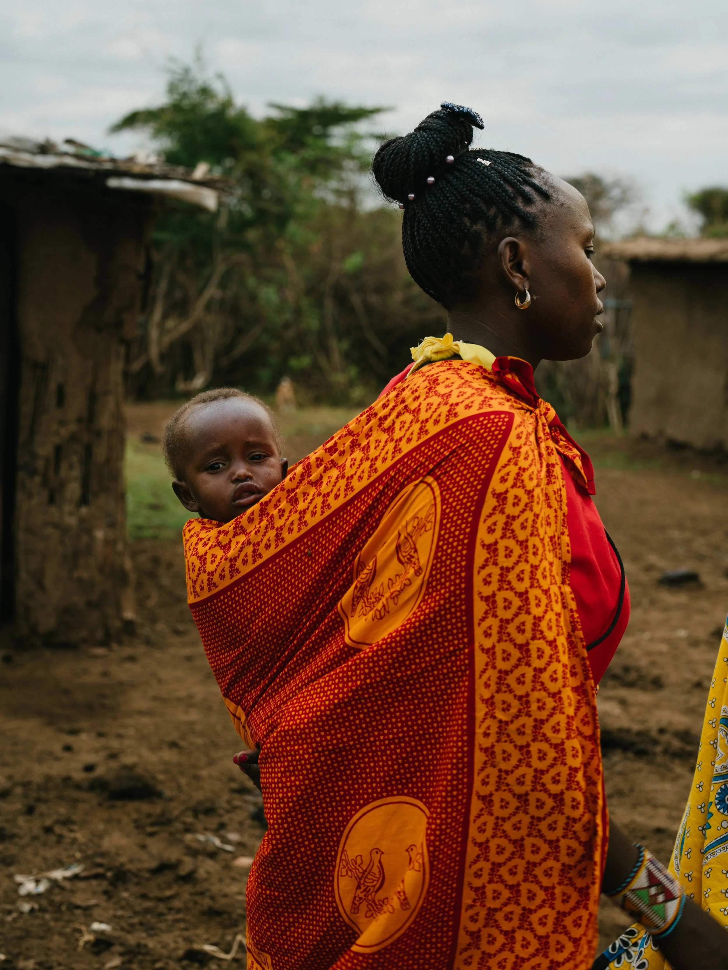 A woman with braided hair and earrings is walking outdoors with a child on her back, wrapped in a bright orange and red patterned cloth. The background shows a rural setting with simple huts and trees.