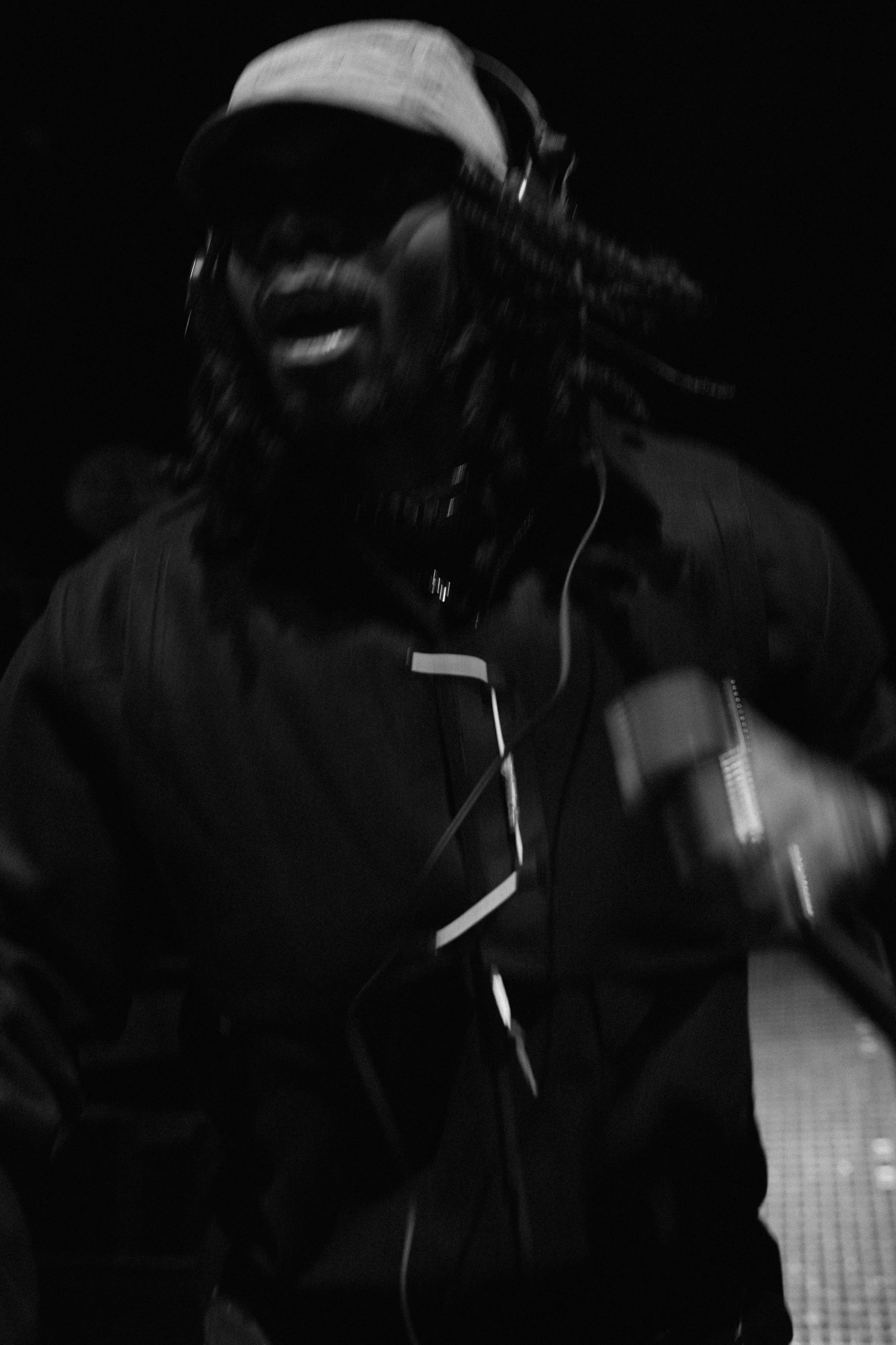 A man with dreadlocks wearing sunglasses, a baseball cap, headphones, and a dark jacket, smiling or singing, holding an object in his hand in a dark setting.