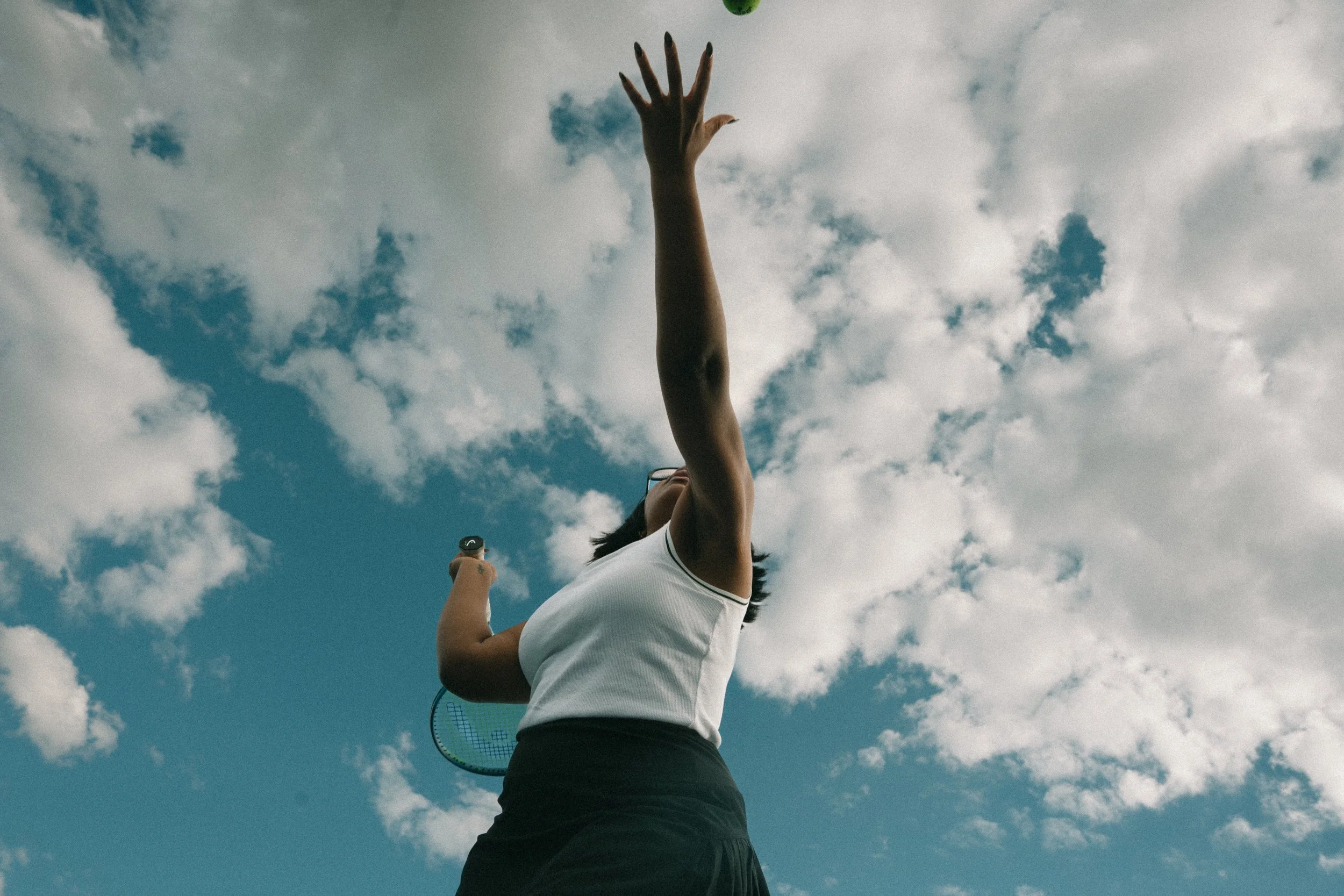 A woman on a tennis court, reaching up with her right hand, holding a tennis racket in her left hand, against a partly cloudy sky.