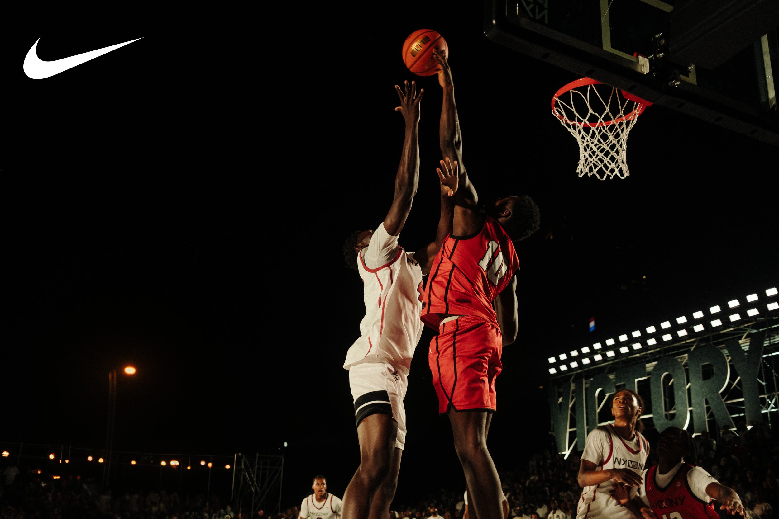Two basketball players are jumping to contest a shot at the basket during a night game; one in a white uniform and the other in a red uniform.